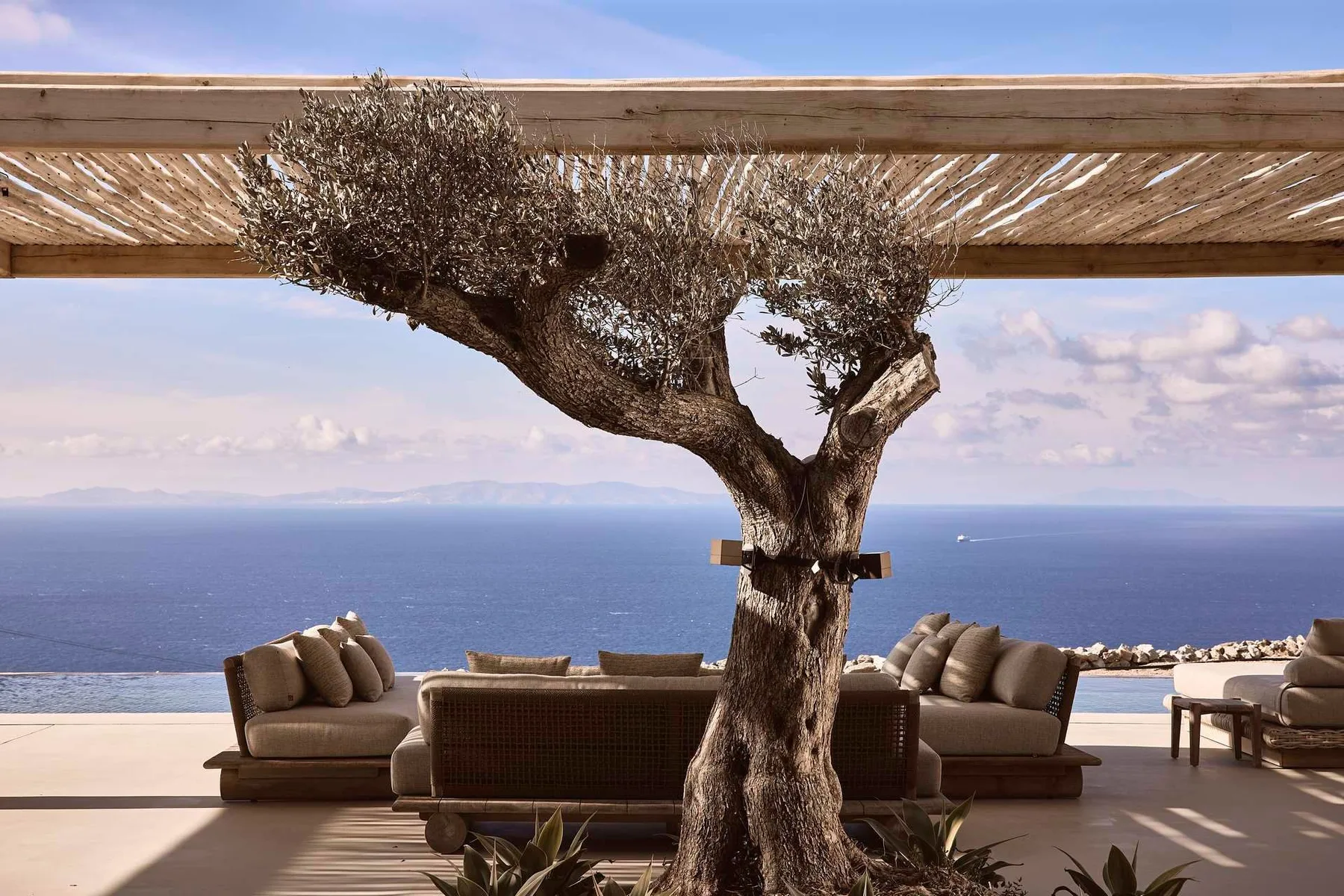 An outdoor seating area overlooks the sea beneath a wooden pergola.