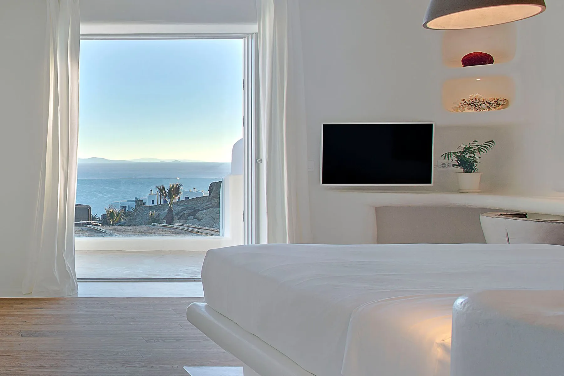 A bedroom with a white bed, television, and a sea view through the window.