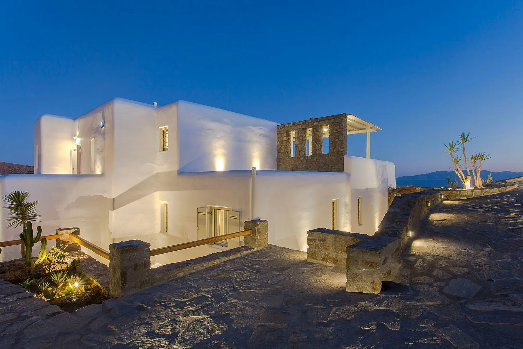 A modern white building is illuminated beside a stone-paved outdoor area at dusk.