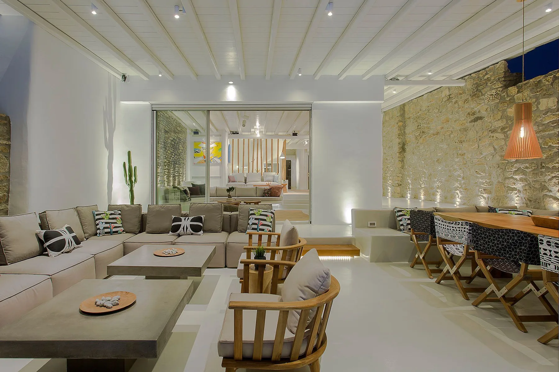 A bright lounge with sectional seating, low tables, and a dining area along a stone wall.