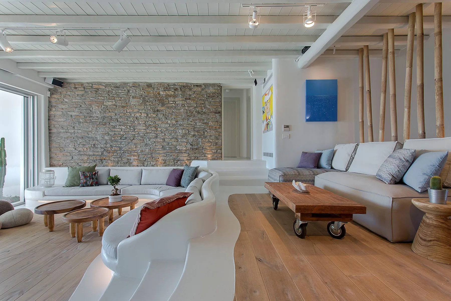 A spacious living room with light seating, wooden tables, and a stone accent wall.