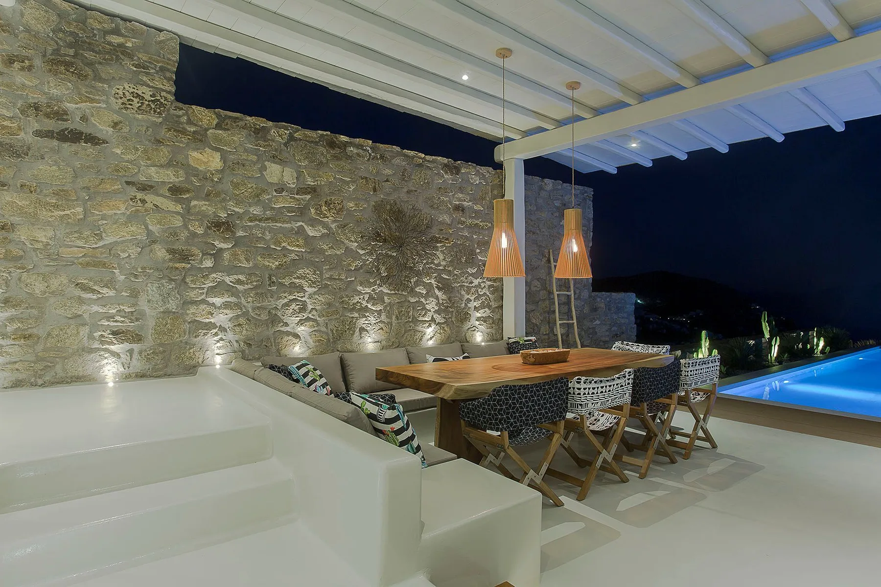 A covered outdoor dining area with a long table beside a pool at night.