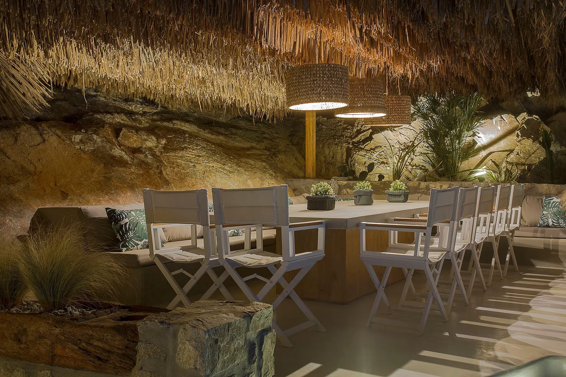 An outdoor dining area with a long table and seating beneath a thatched canopy.