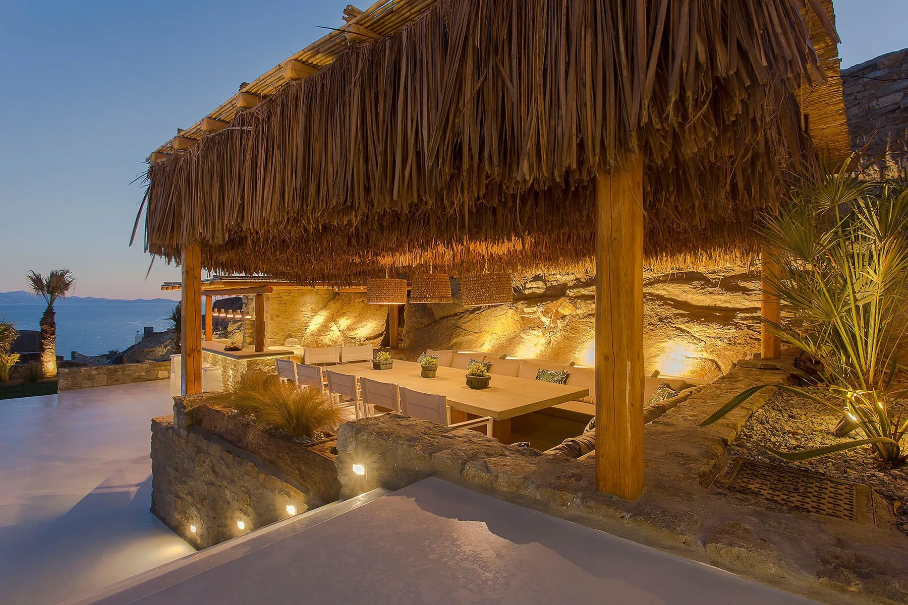 An outdoor dining area sits beneath a thatched canopy beside the sea.
