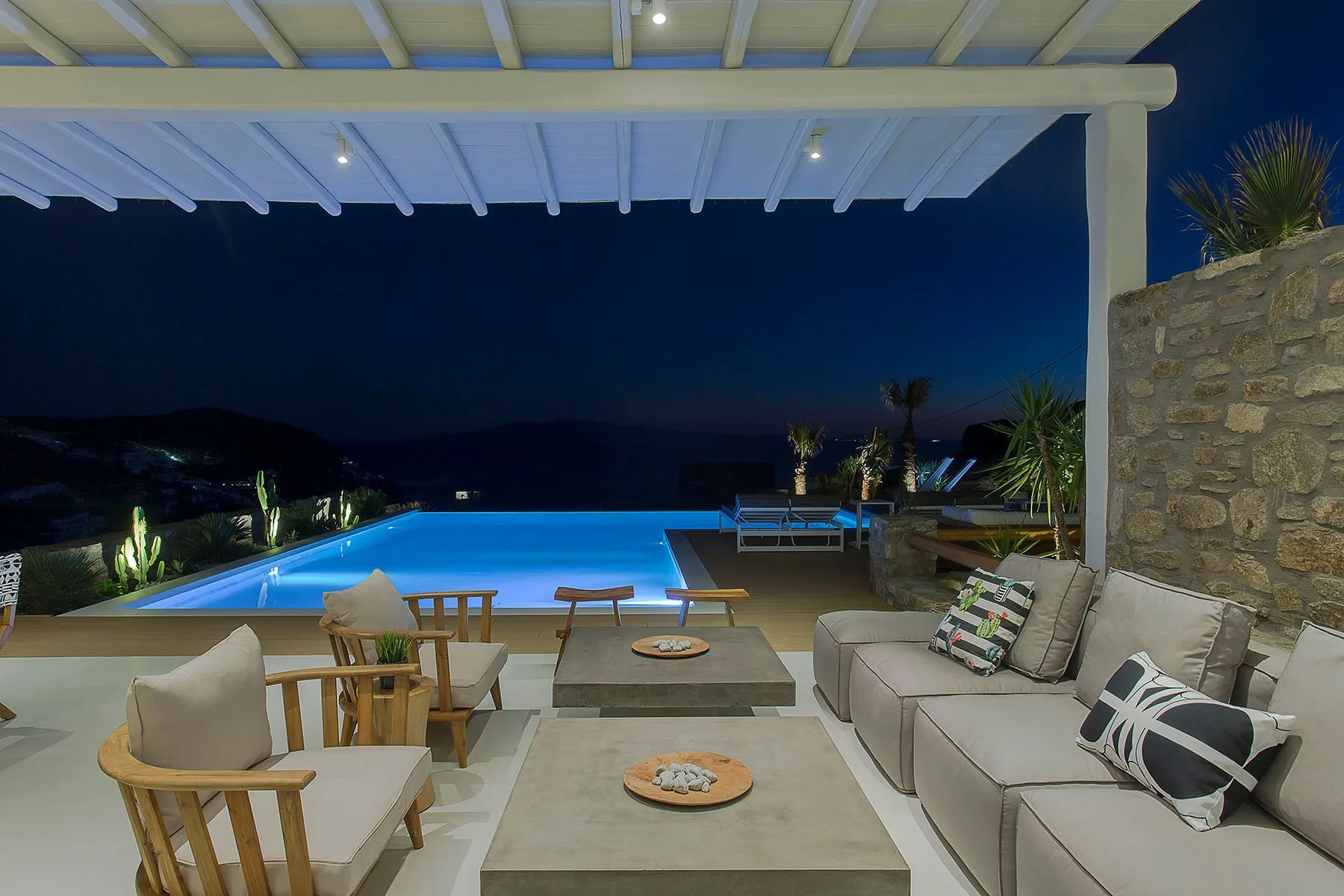 A covered outdoor lounge and dining area overlooks a brightly lit swimming pool at night.