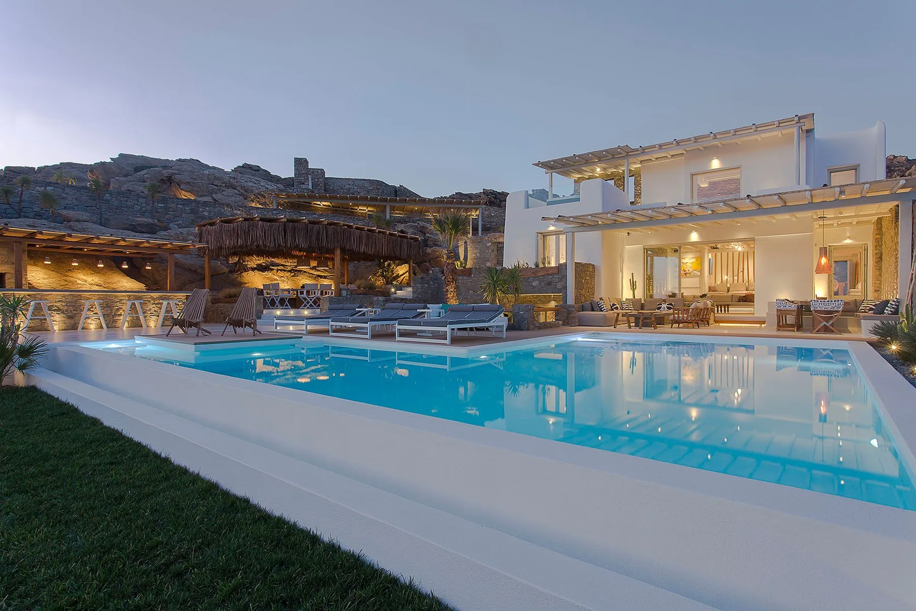 A lit outdoor swimming pool sits beside a modern house at dusk.