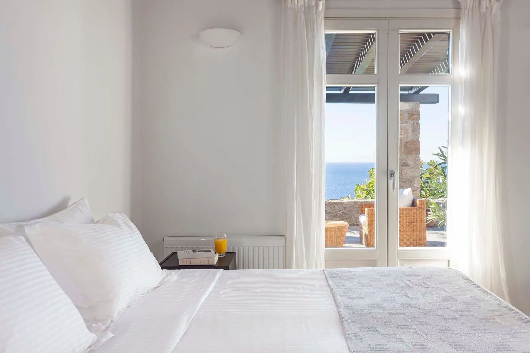 A bright bedroom with white bedding and a glass door opening toward a sea view.