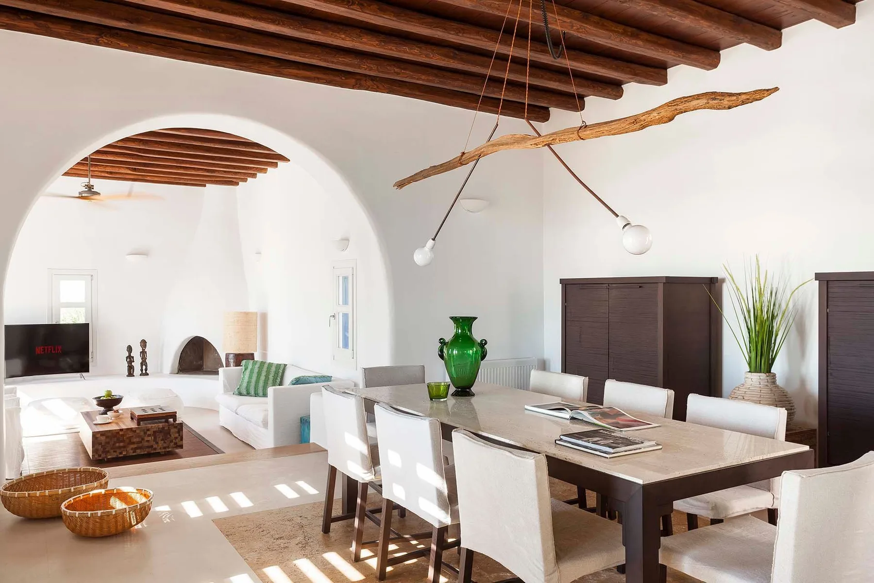 A bright dining and living area features a long table beneath exposed wooden ceiling beams.
