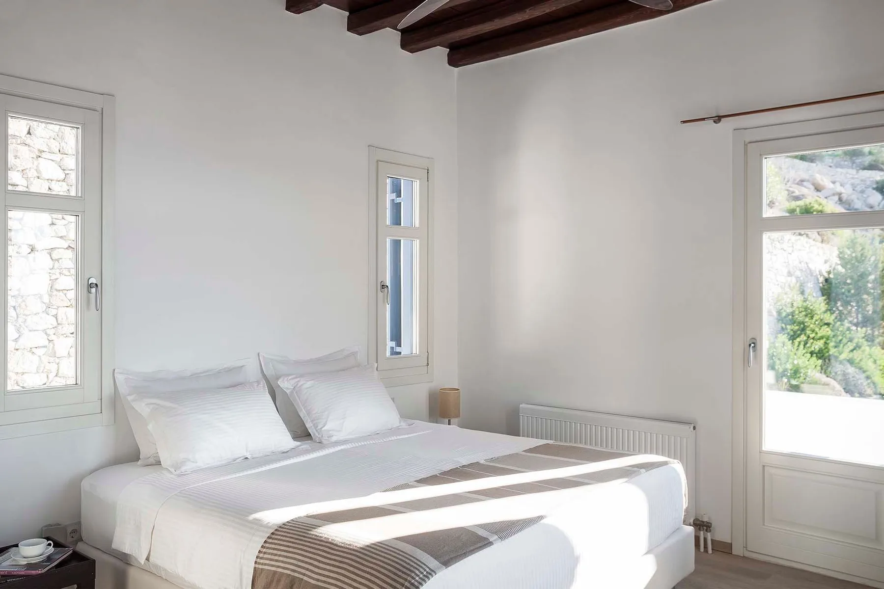 A bright bedroom with a neatly made bed beneath exposed wooden ceiling beams.