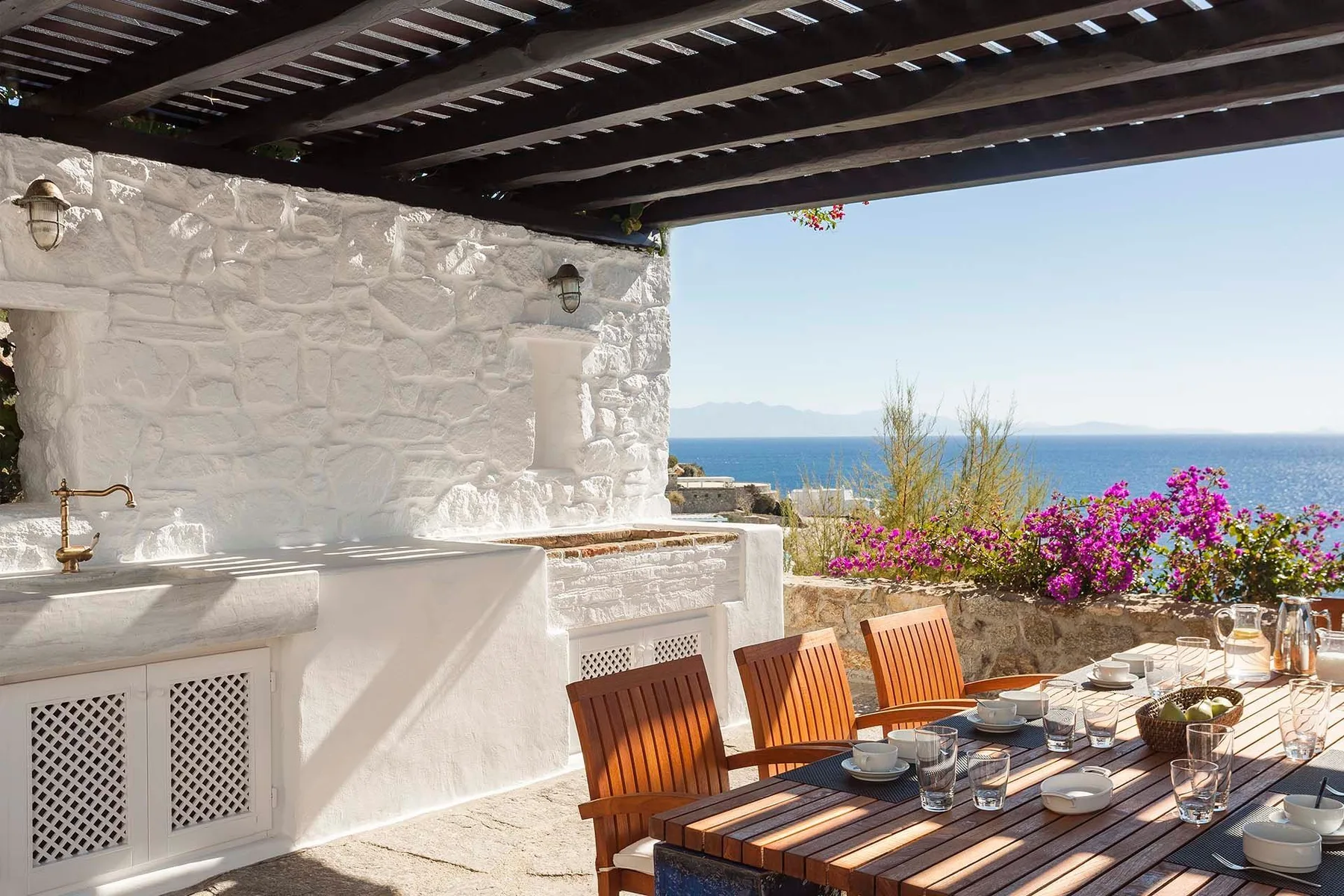 A seaside dining patio with a wooden table beneath a shaded pergola.