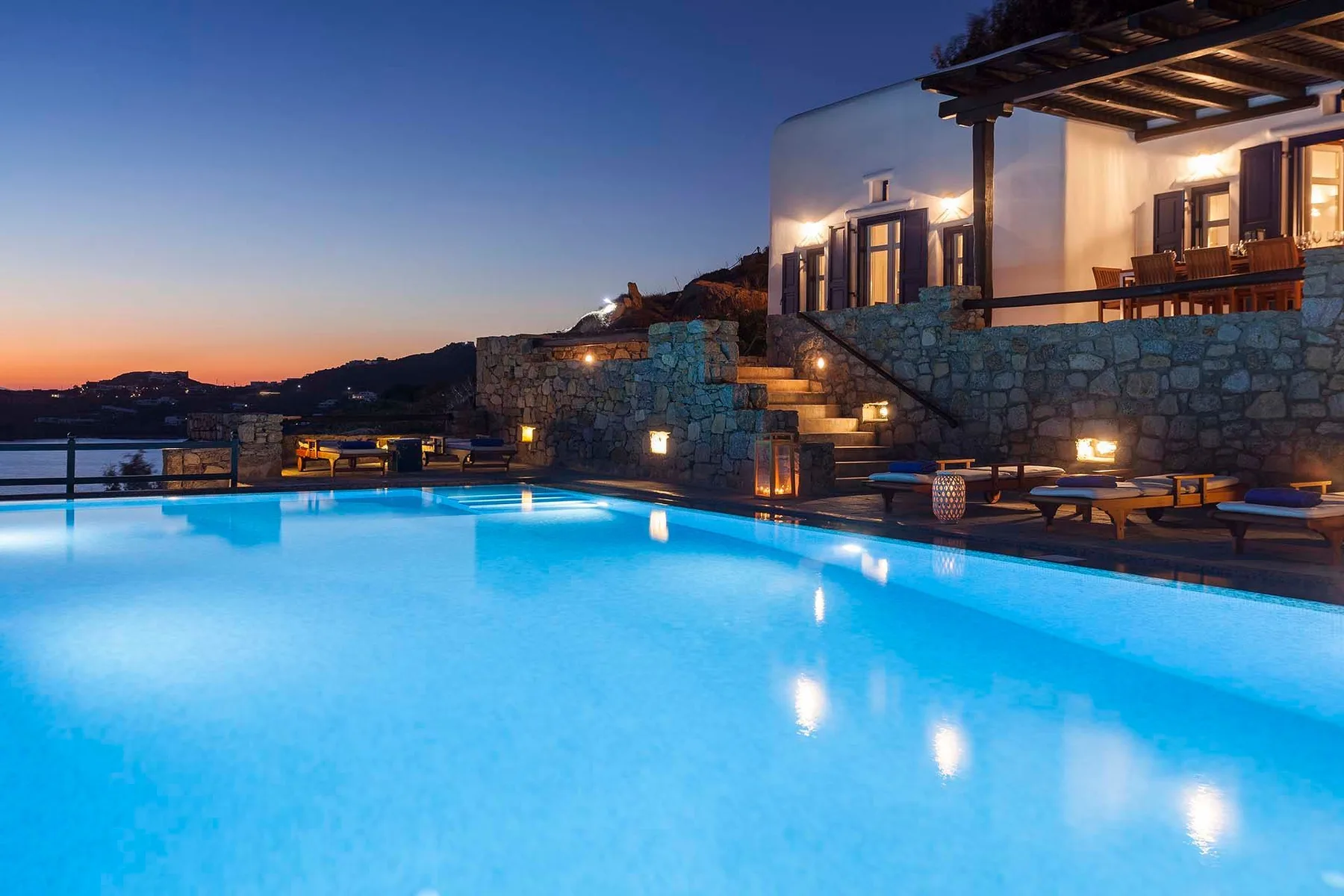 An illuminated swimming pool sits beside a stone terrace at dusk.