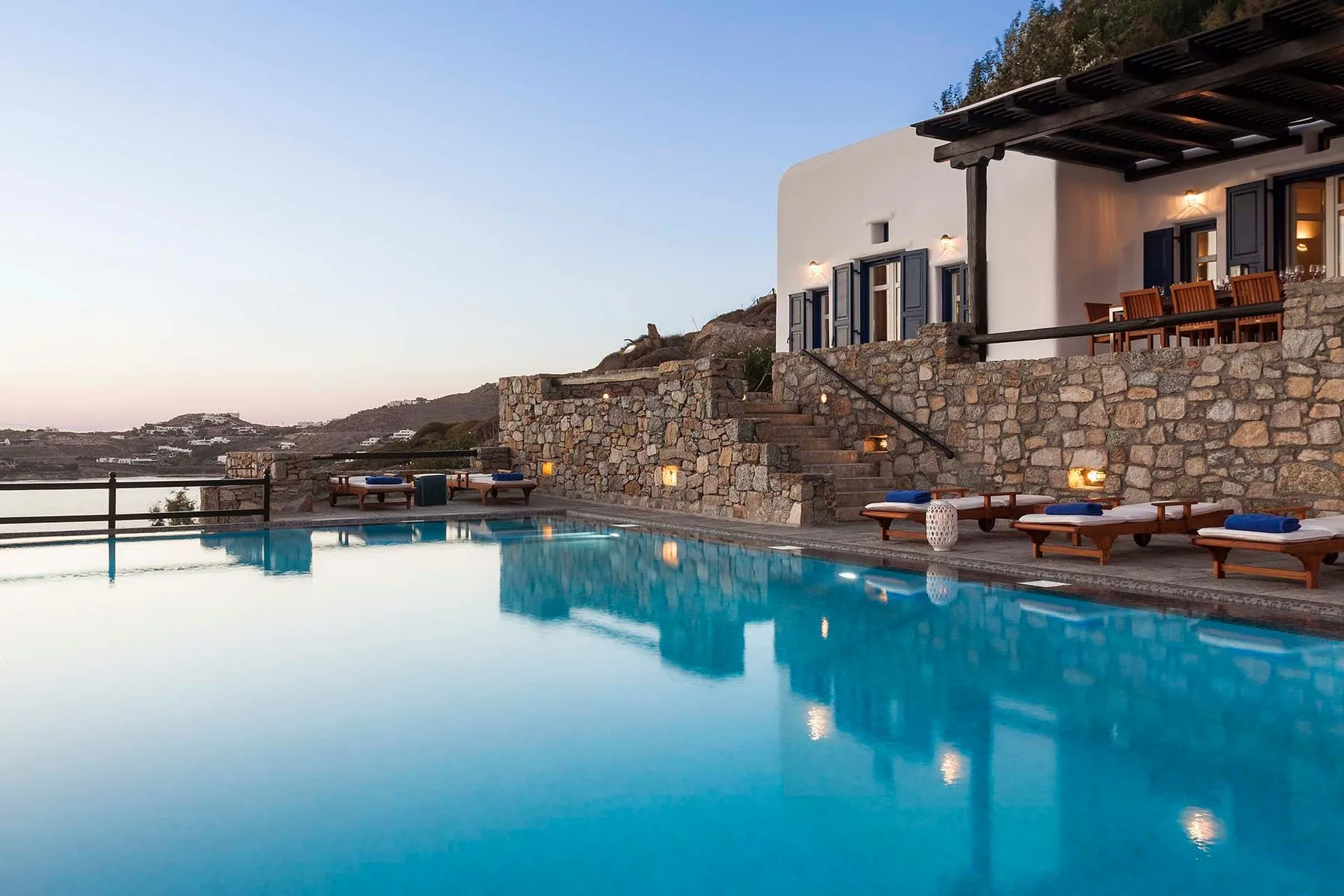 An outdoor pool beside a stone terrace and white building at dusk.
