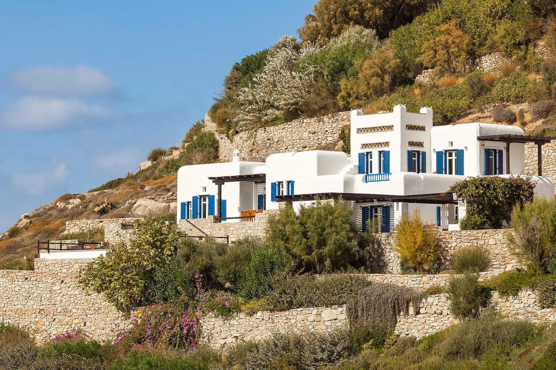 White hillside buildings with blue shutters sit among stone walls and vegetation.