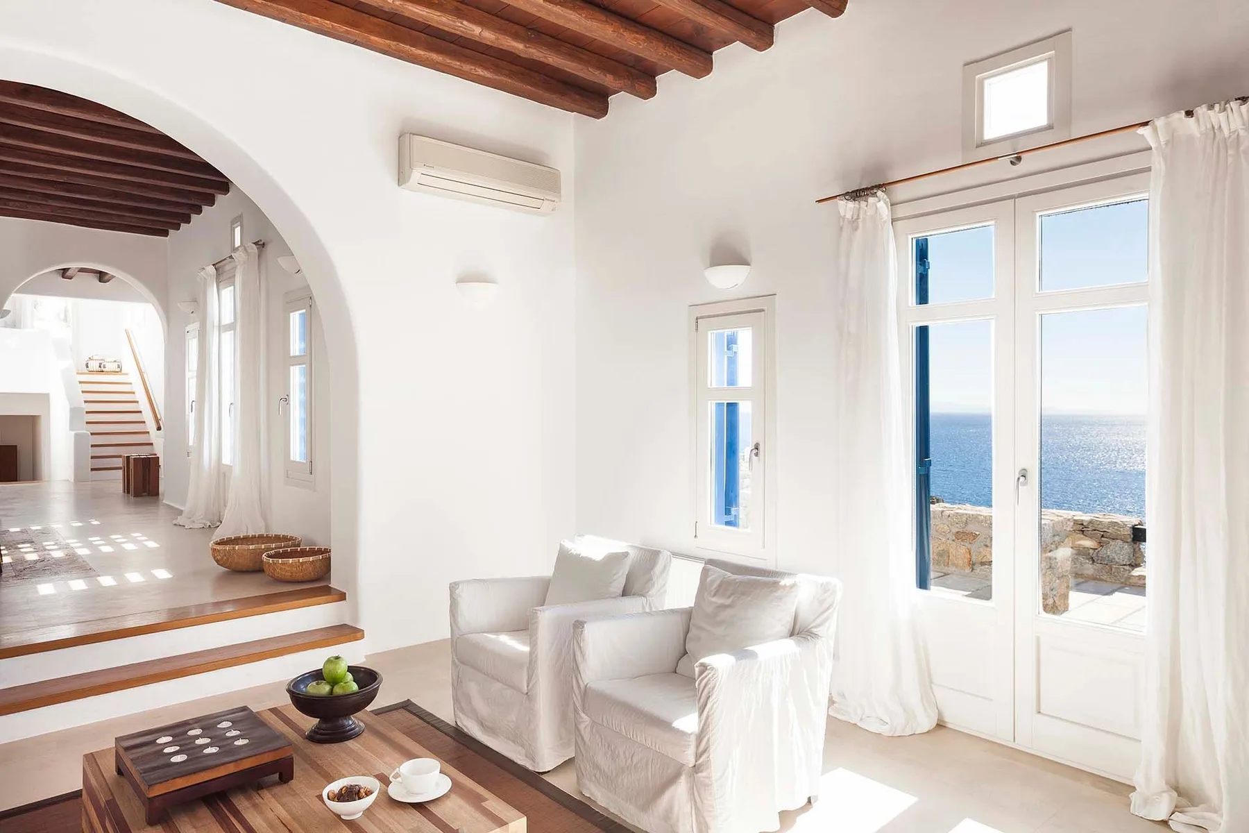 A bright living room with white seating beside open doors overlooking the sea.