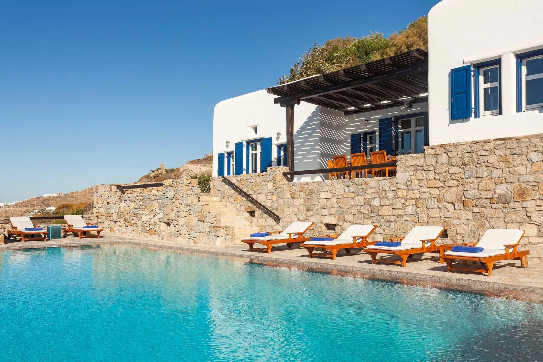A swimming pool sits beside a white building with blue shutters and outdoor seating.