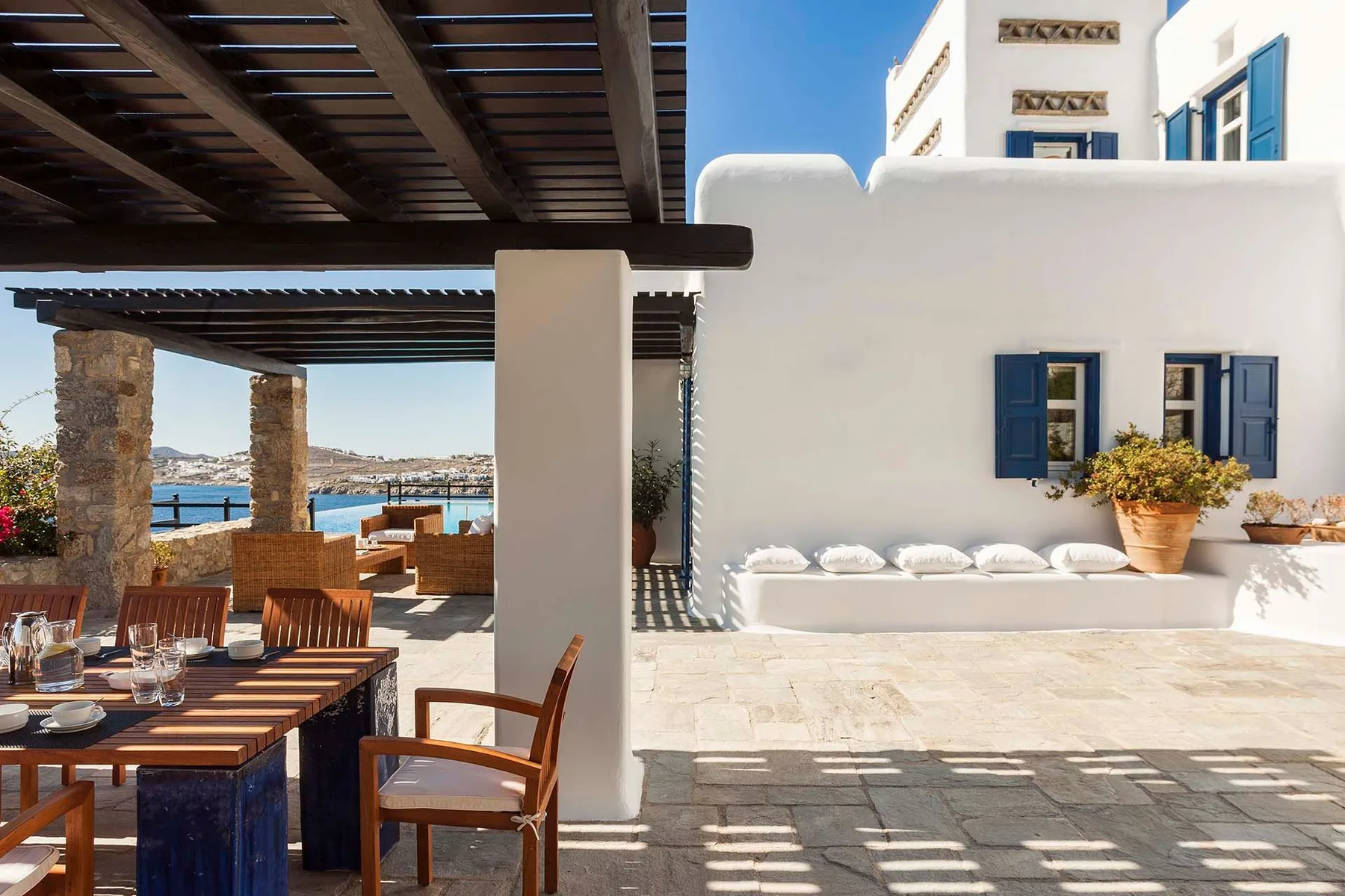 A shaded outdoor dining terrace beside a white building, with a view of the sea.