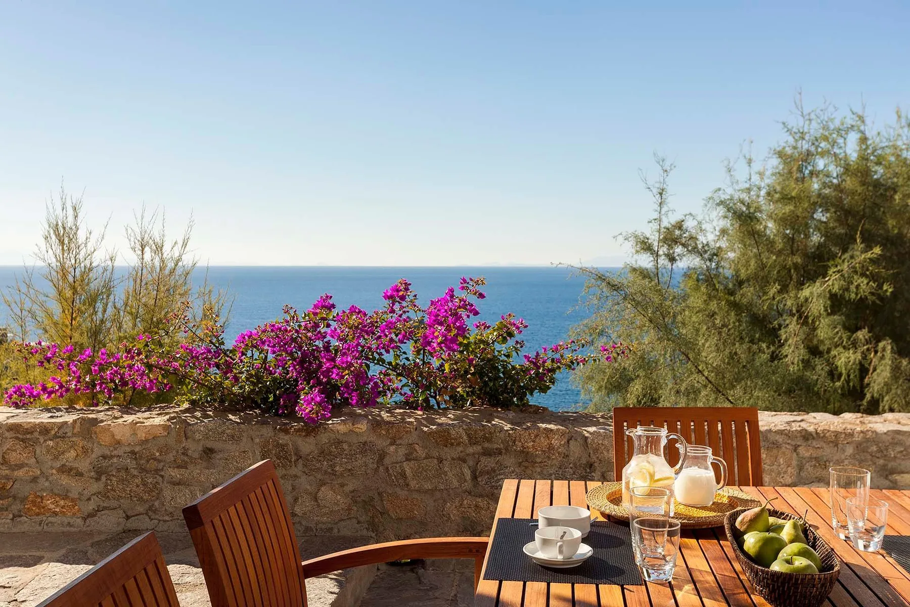 An outdoor dining area overlooks the sea, with a wooden table set with dishes and fruit.