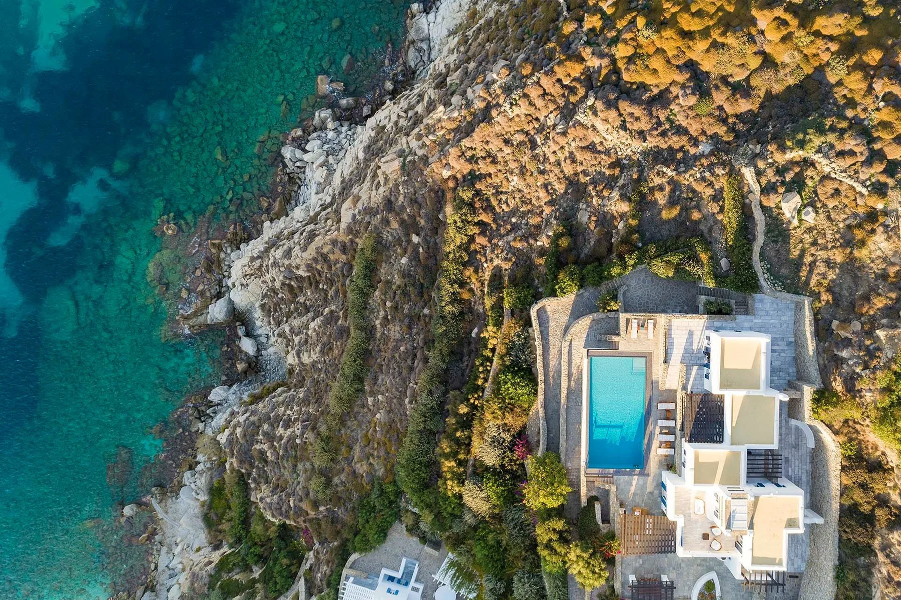 An aerial view shows a seaside home with a swimming pool beside rocky turquoise water.