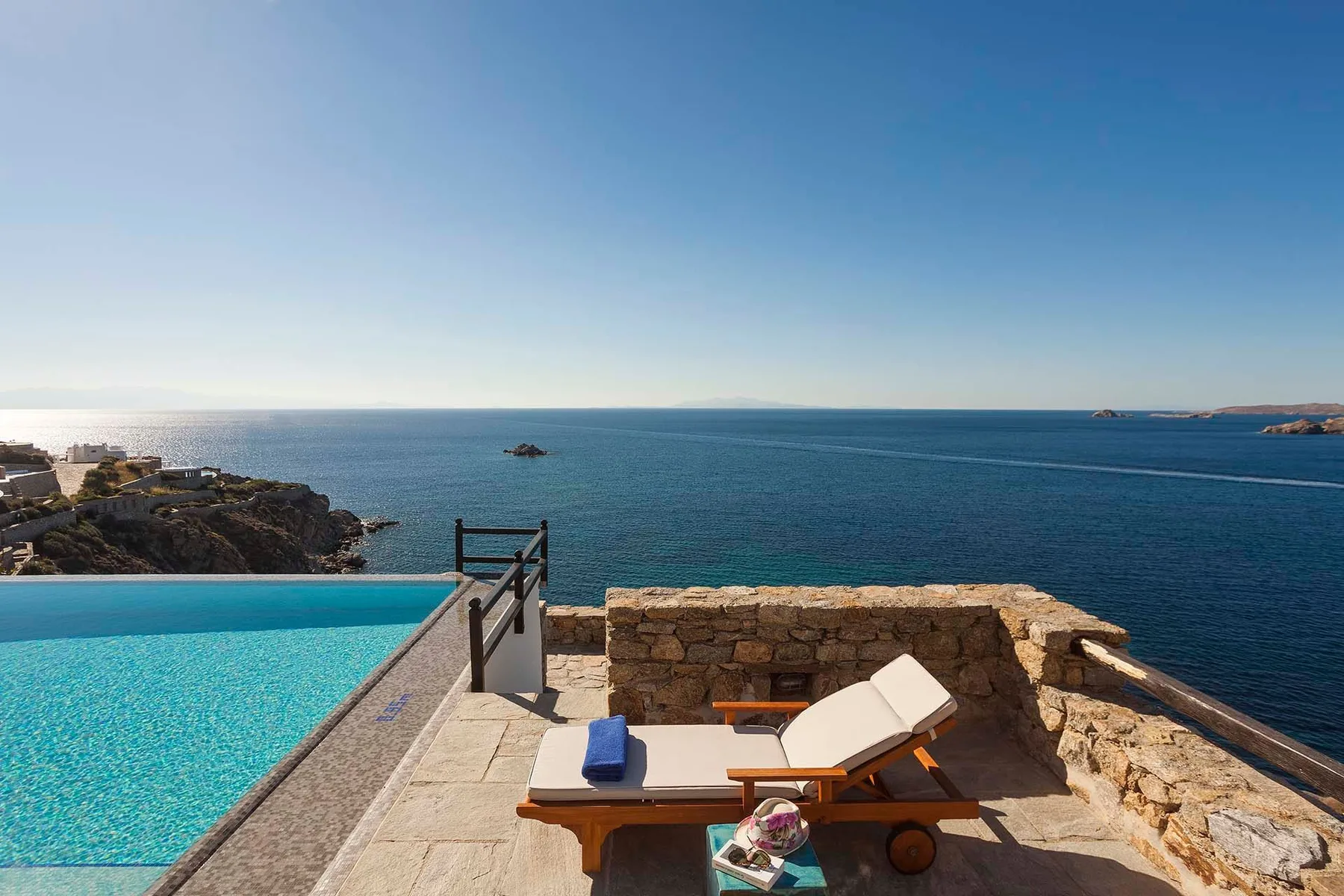 A seaside pool terrace overlooks the open water, with a cushioned sun lounger nearby.