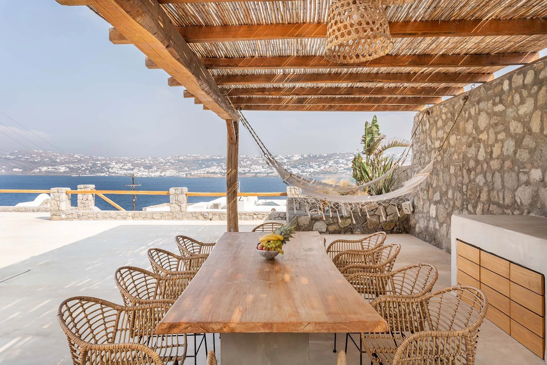 A shaded outdoor dining area with a long table and seating overlooks the sea.