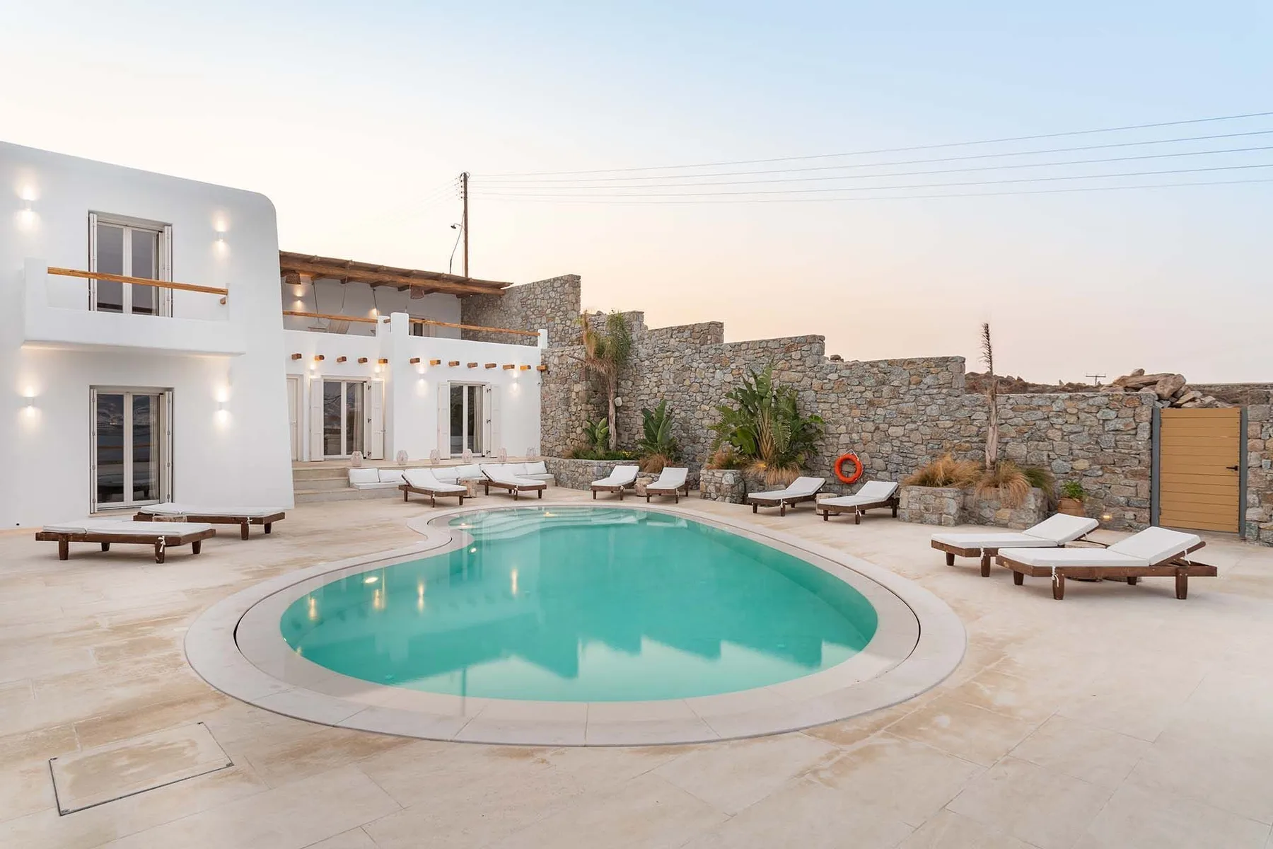 A turquoise pool sits in a paved courtyard beside a white building and stone wall.