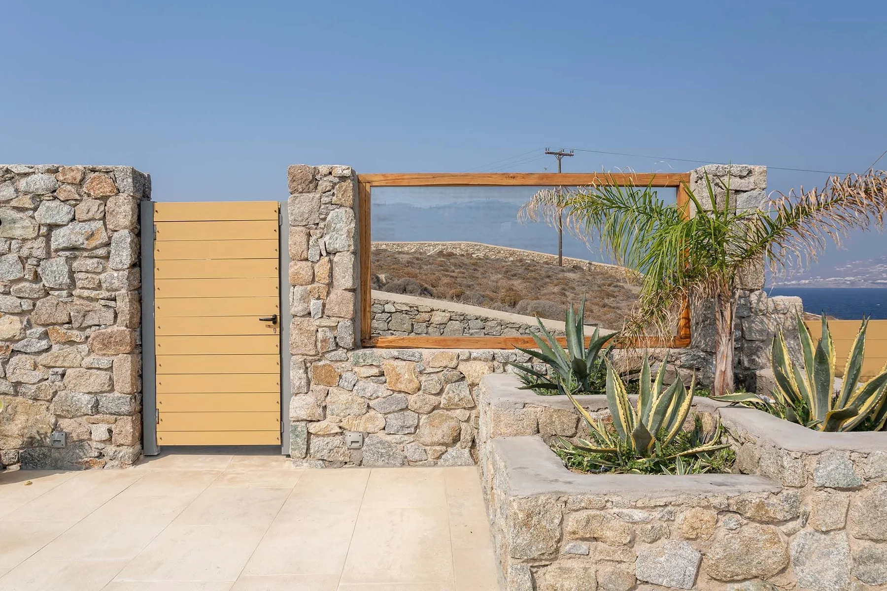 A stone garden entrance with a wooden gate, framed view, and desert plants.