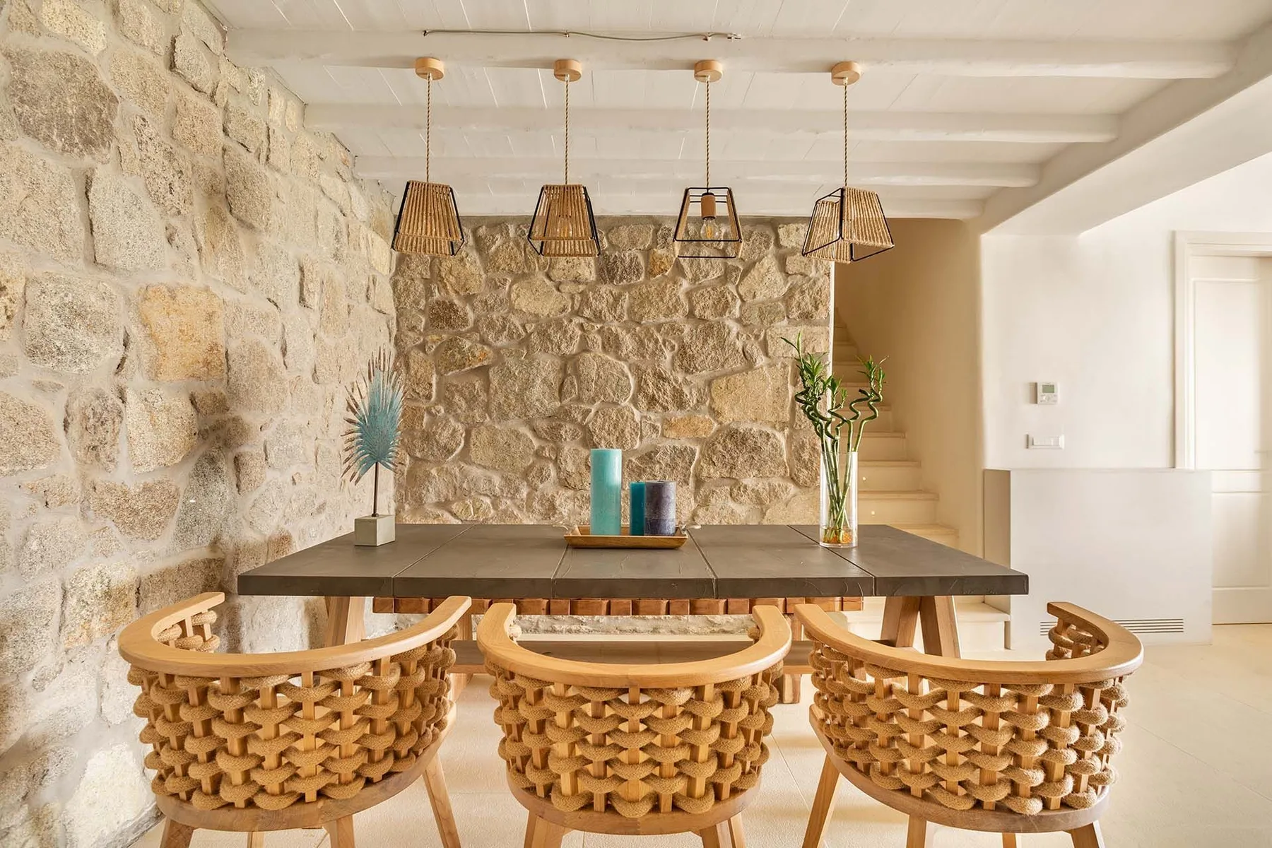 A dining area with a long wooden table and woven chairs beside a stone wall.