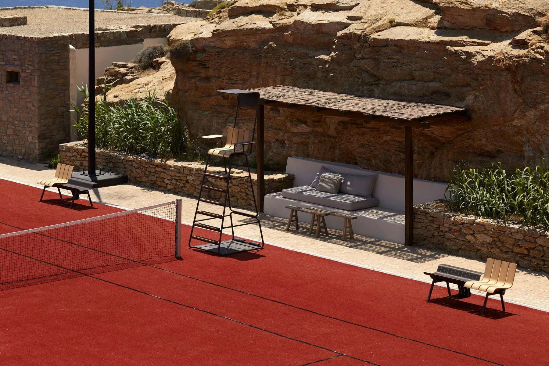 An outdoor tennis court with seating beneath a shaded pergola.