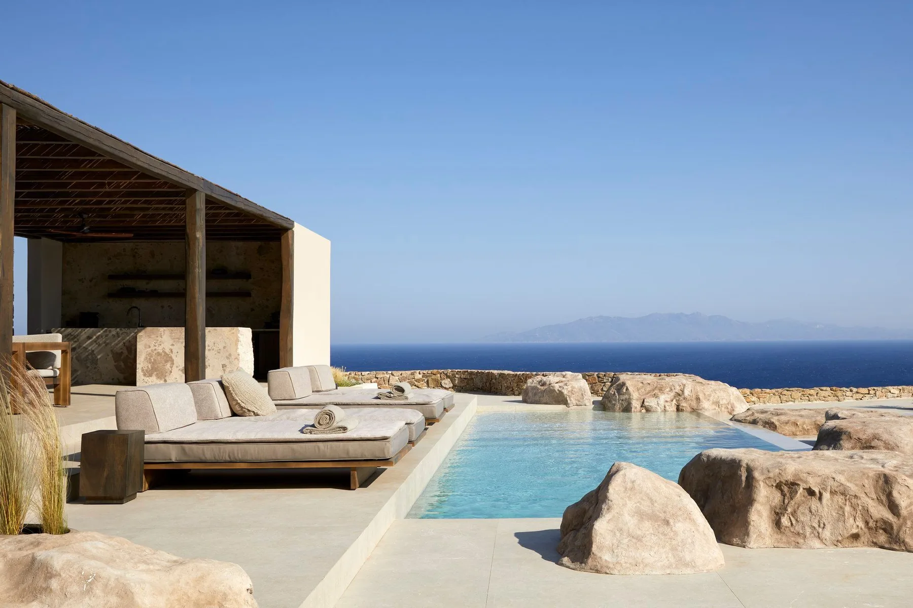 An outdoor pool beside a shaded seating area, with rocky terrain and the sea beyond.