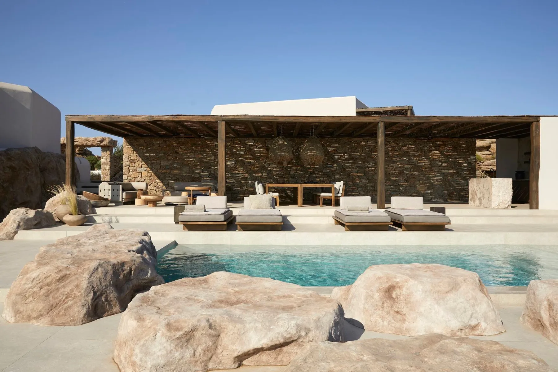 An outdoor pool sits in front of a shaded seating area and stone wall.