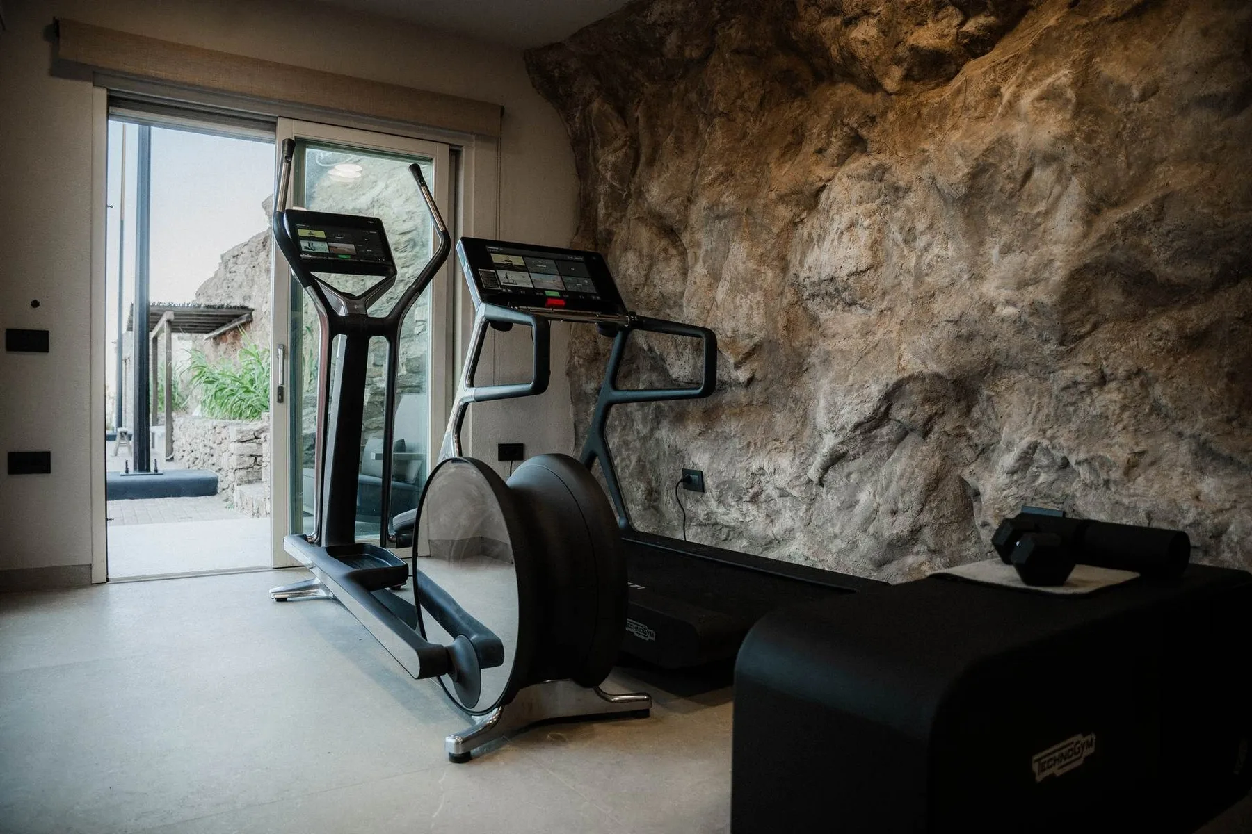 A small gym with cardio exercise machines beside a natural stone wall.