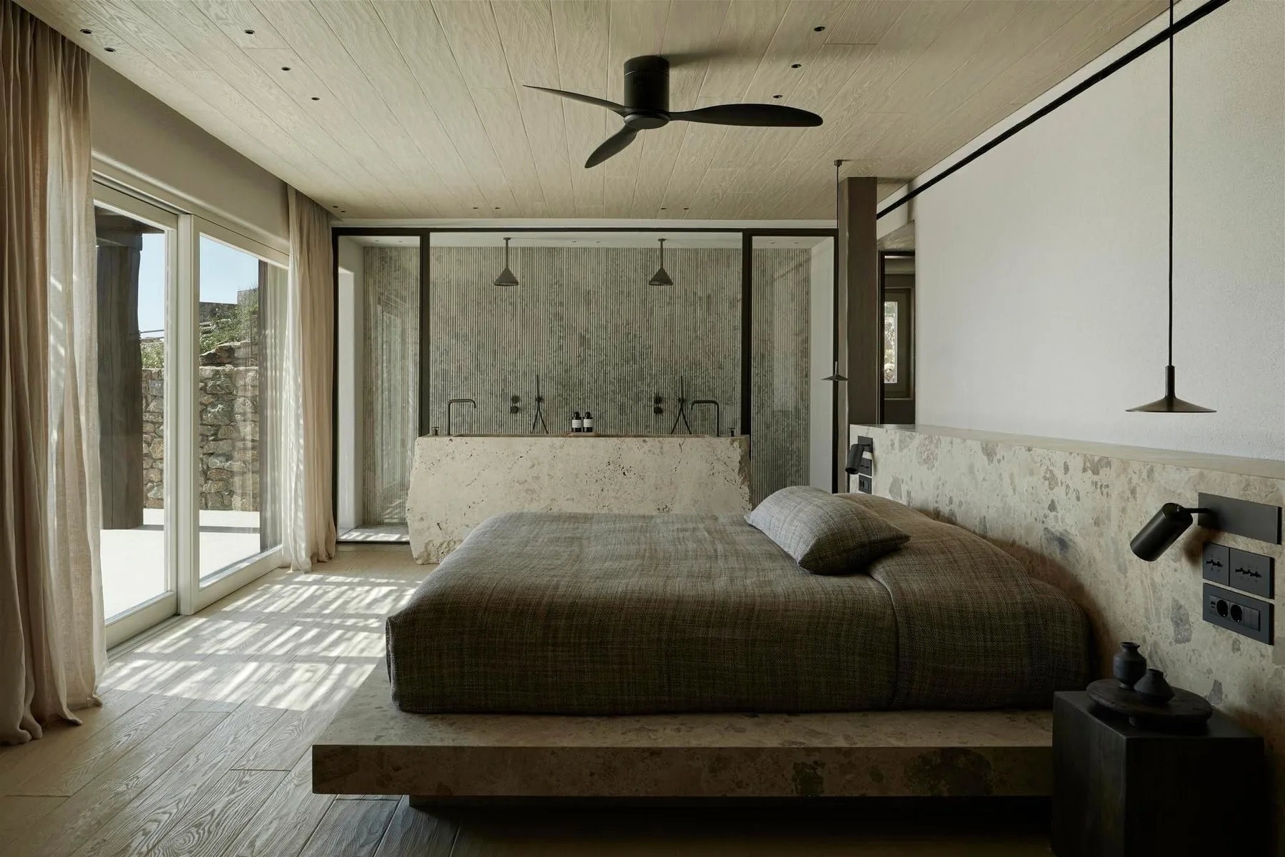 A bedroom with a low bed beside large glass doors and a stone wall.