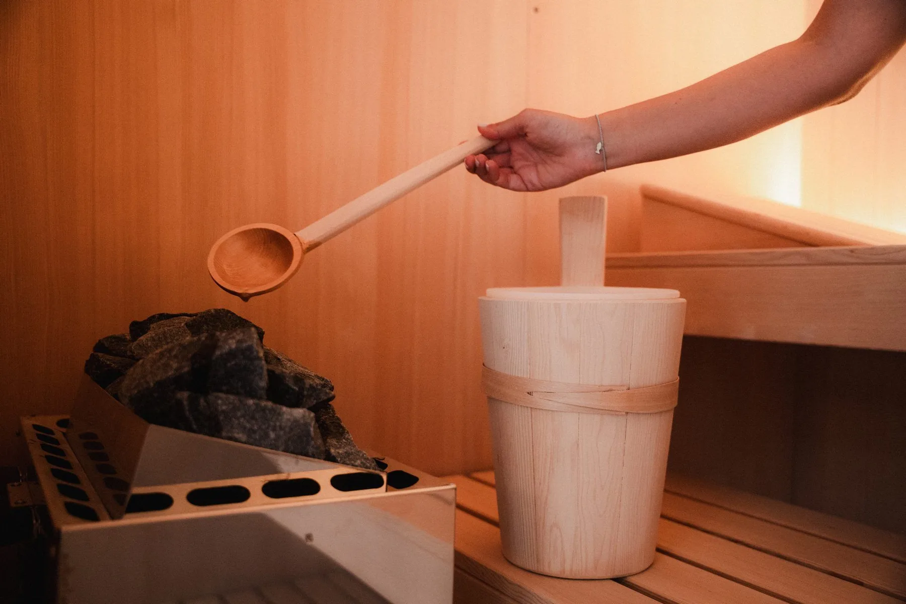 A sauna heater with stones, a wooden bucket, and a ladle in a wood-paneled sauna.