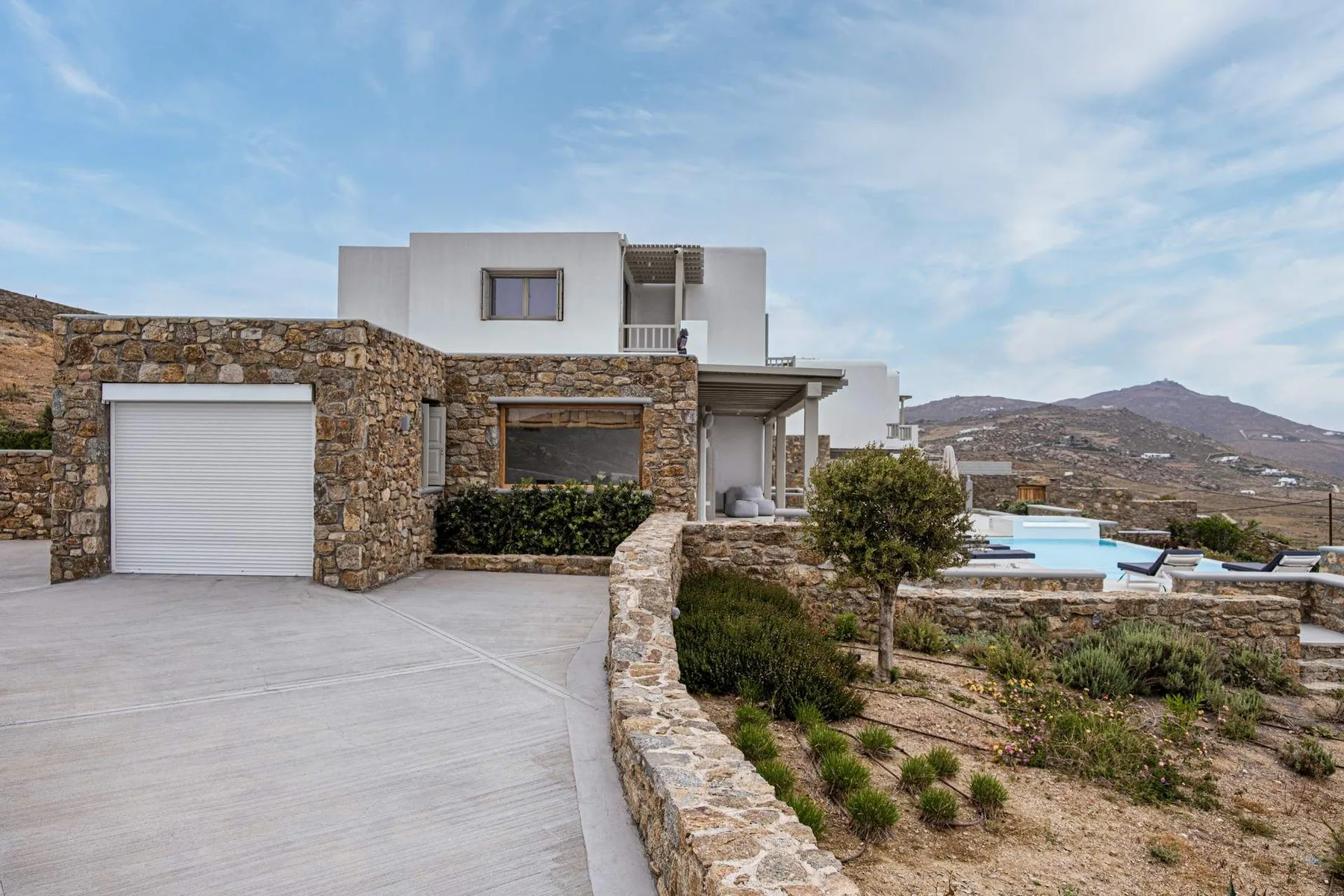A modern house with stone walls, a driveway, and an outdoor pool in a hilly landscape.