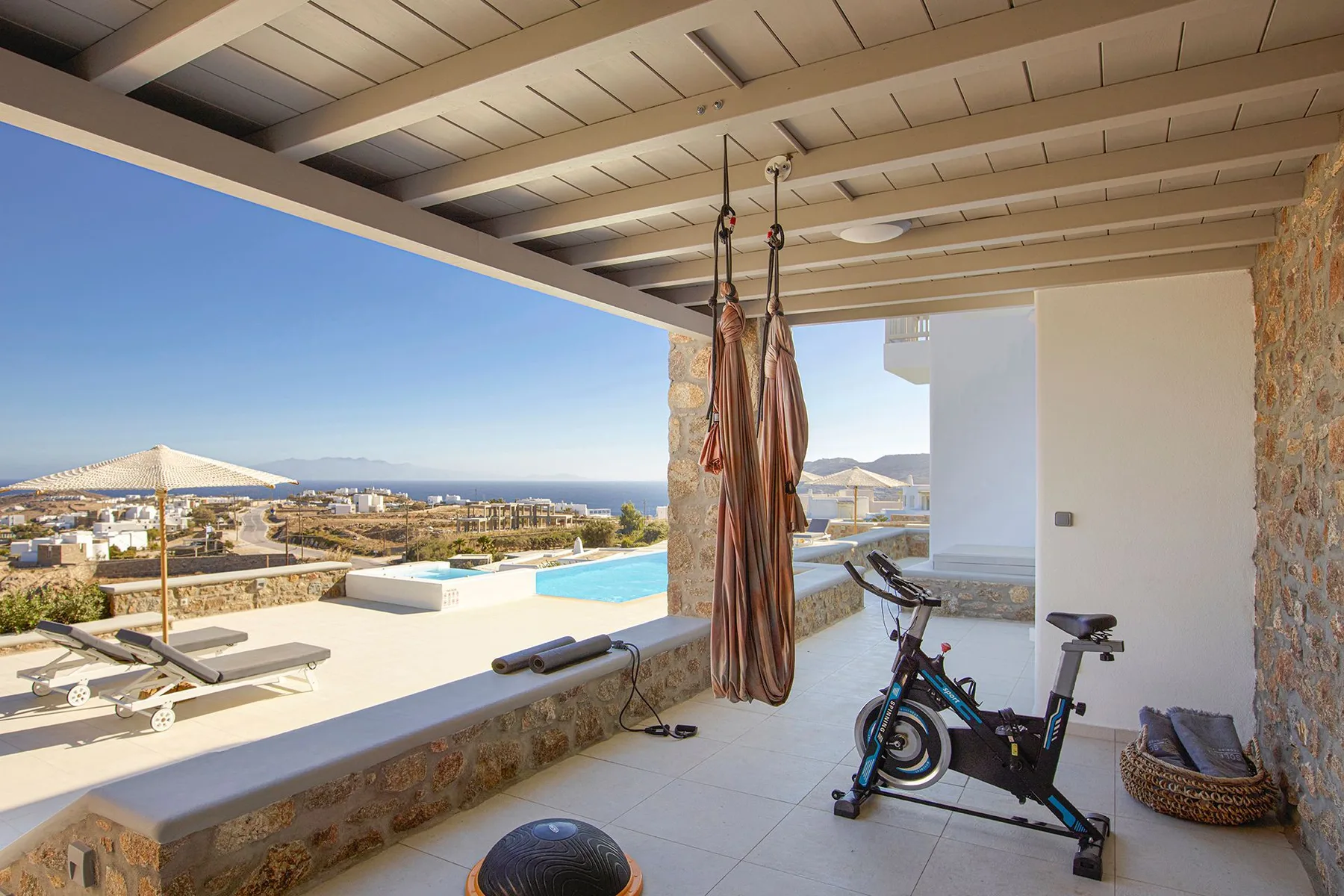 A covered terrace overlooks a swimming pool and the sea, with exercise equipment nearby.