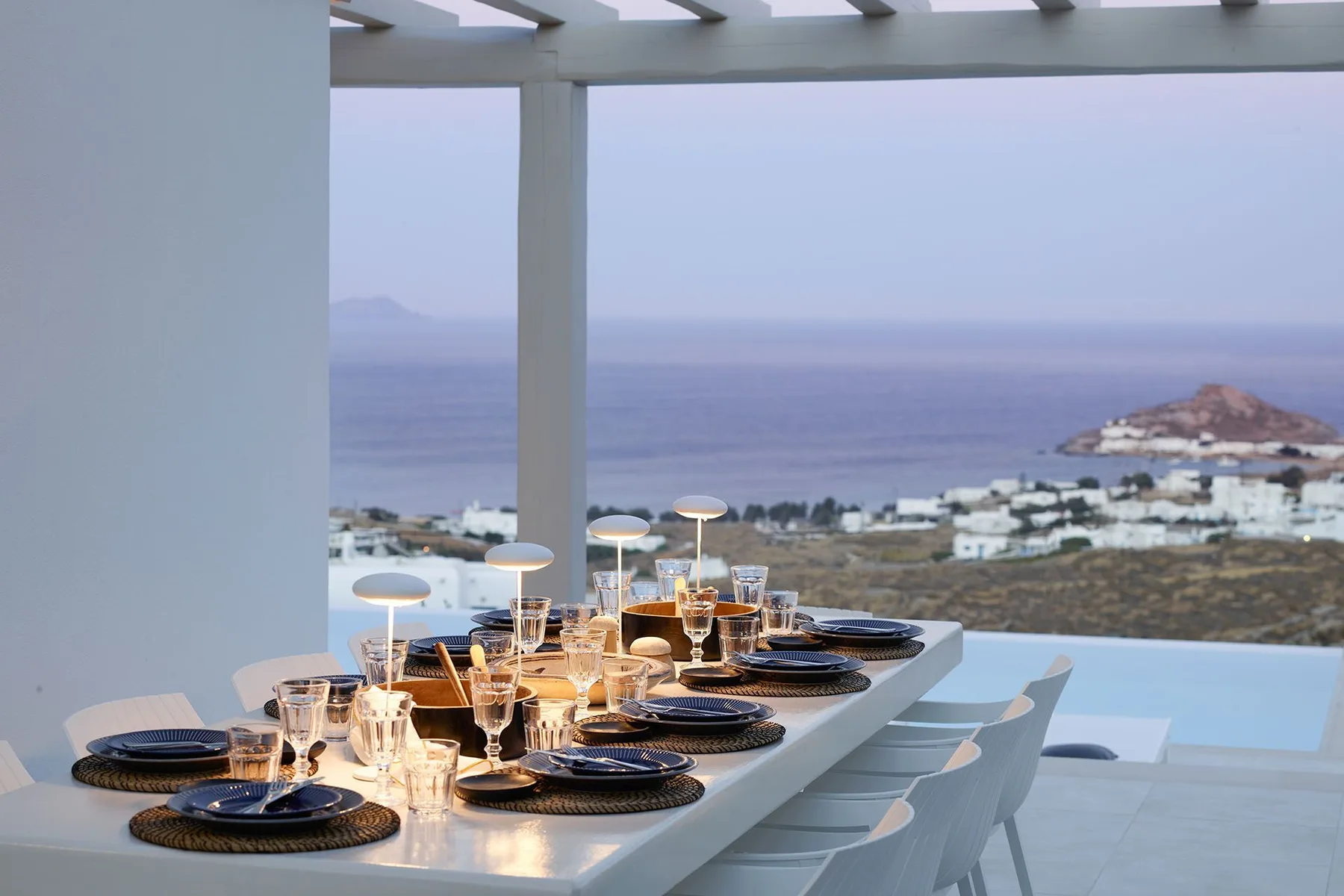 A dining table set for a meal overlooks the sea and a coastal settlement.