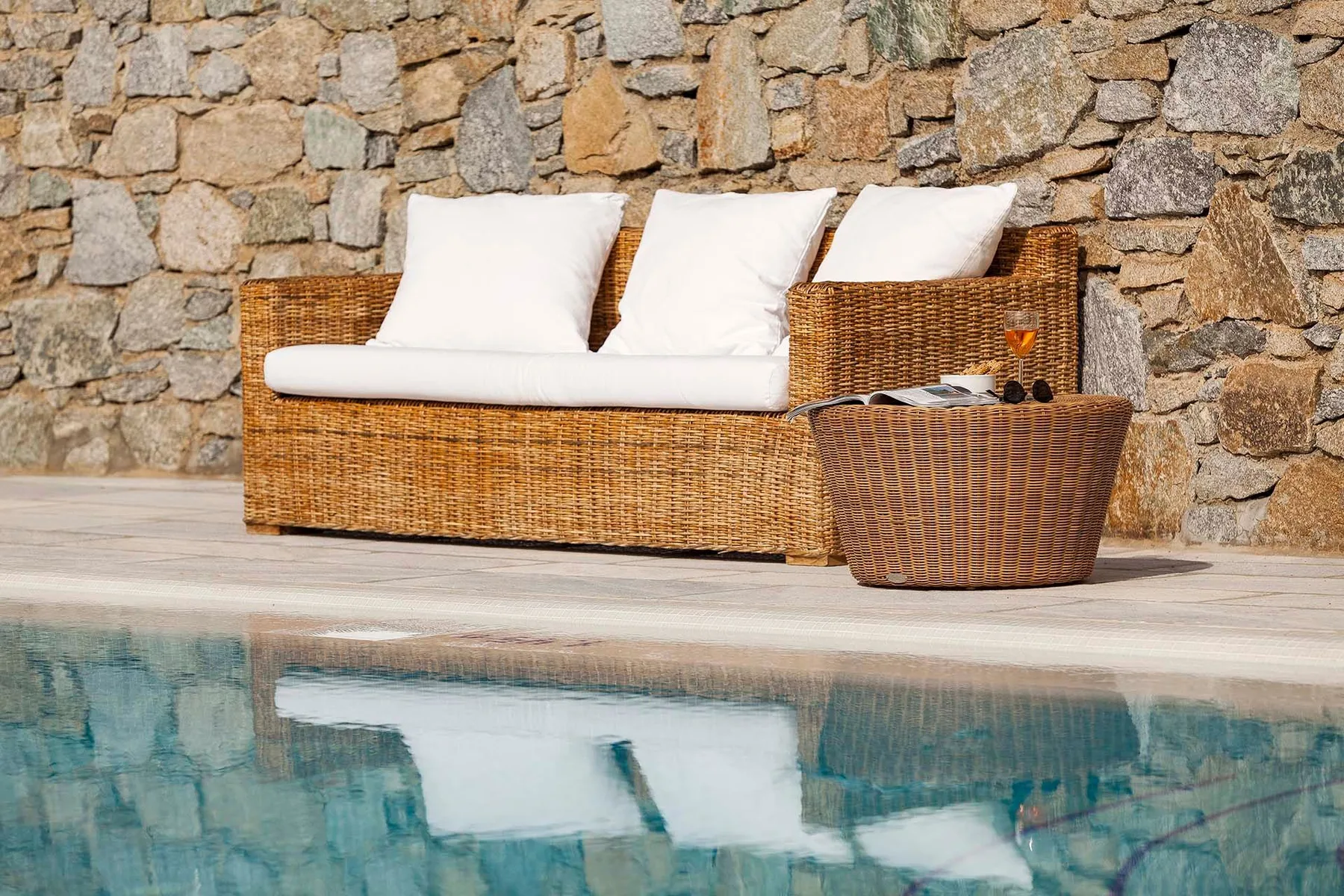 Una zona de asientos de mimbre con cojines junto a una piscina y un muro de piedra.