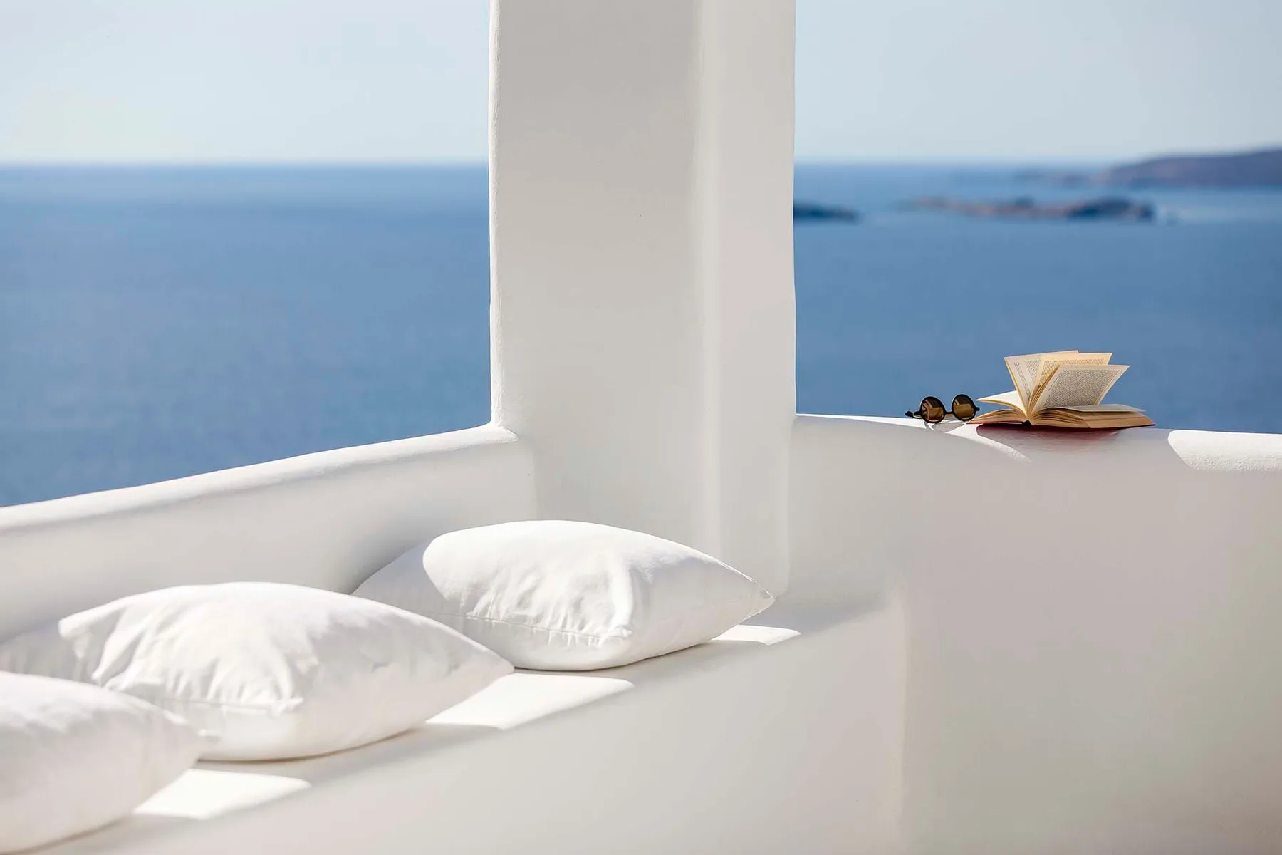 Una zona de asiento acolchada blanca da al mar, con un libro abierto sobre el borde.