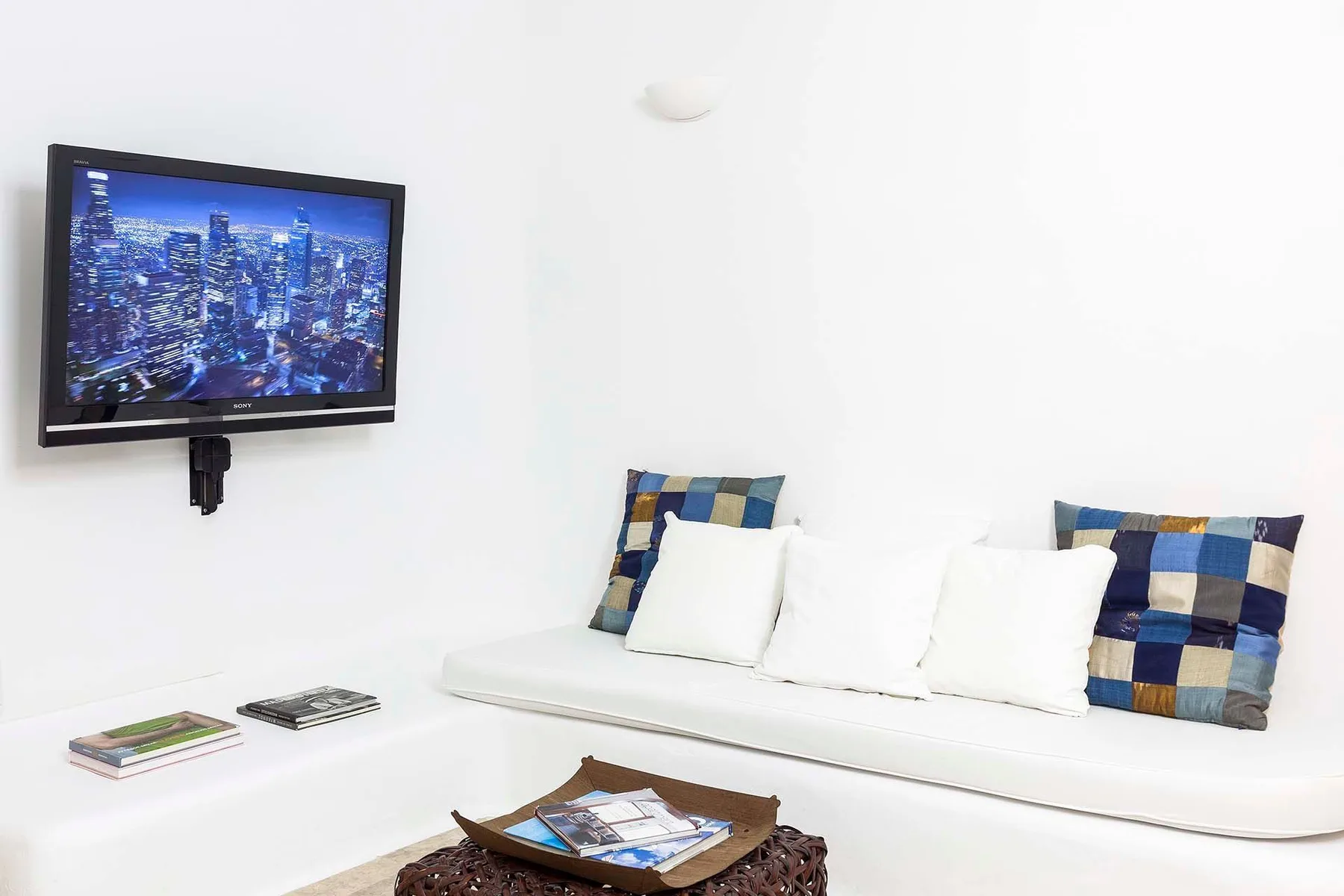 Una sala luminosa tiene un sofá blanco con cojines coloridos y un televisor en la pared.