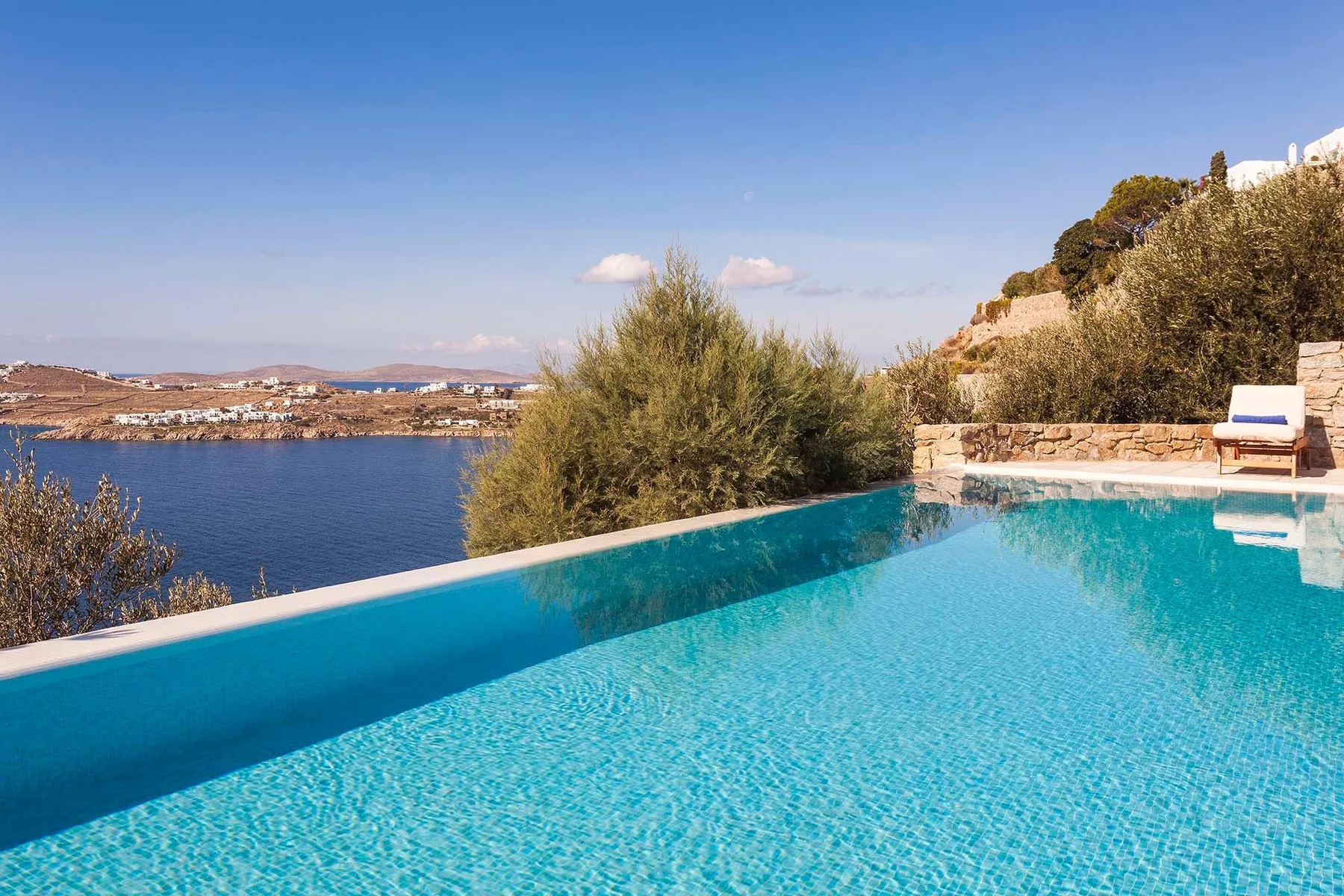 Una piscina infinita da al mar azul, con una tumbona en la terraza de piedra.