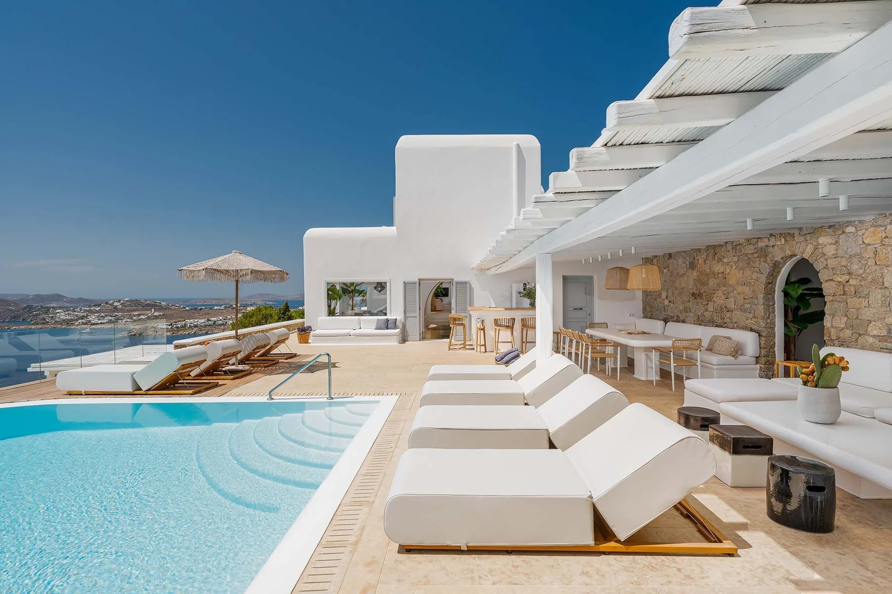 A sunny poolside terrace with white loungers, outdoor seating, and a sea view.
