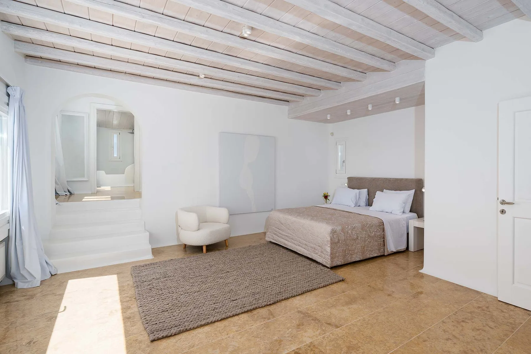 A bright bedroom with a bed, an area rug, and exposed ceiling beams.