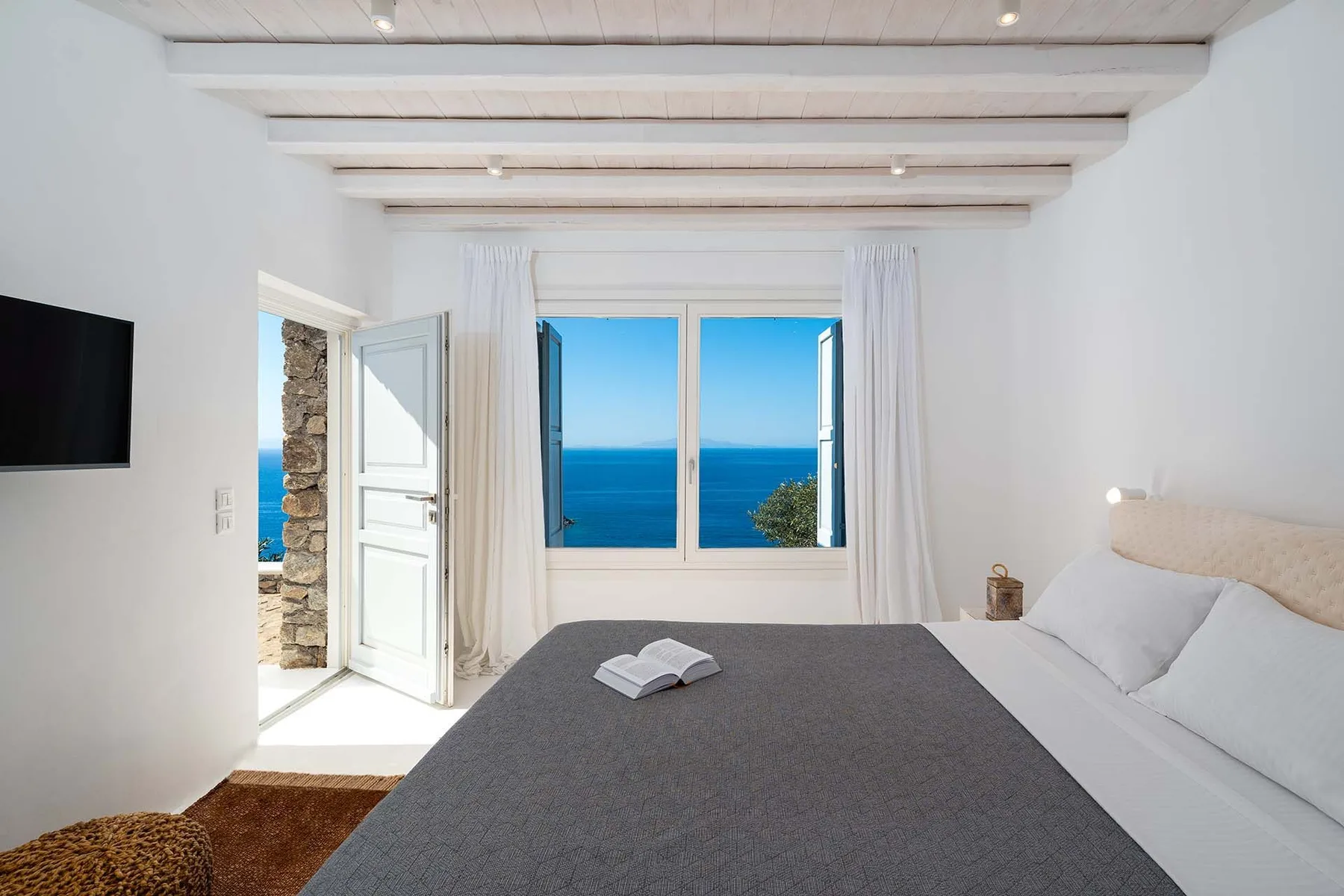 A bedroom with a bed beside an open window overlooking the sea.
