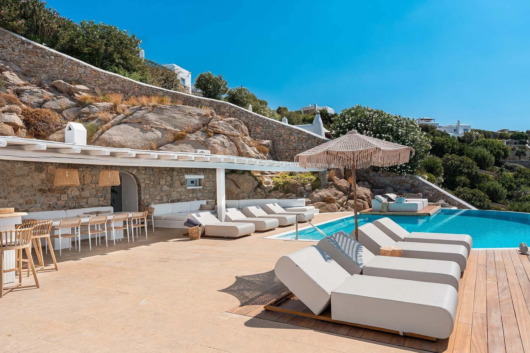 A sunlit pool terrace with lounge seating, dining tables, and a stone wall.