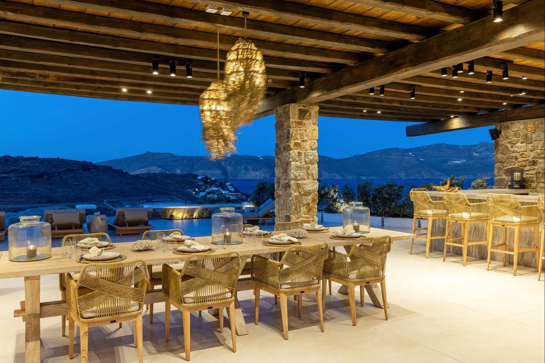 An open-air dining area with a long table and woven seating overlooks a mountain landscape.