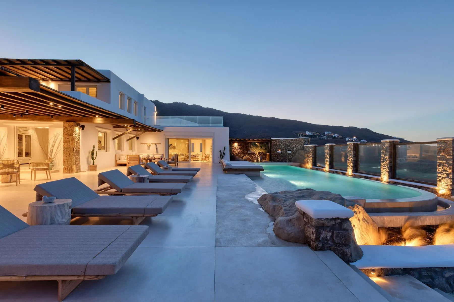 A rooftop terrace with lounge seating overlooks a lit swimming pool and distant hills.