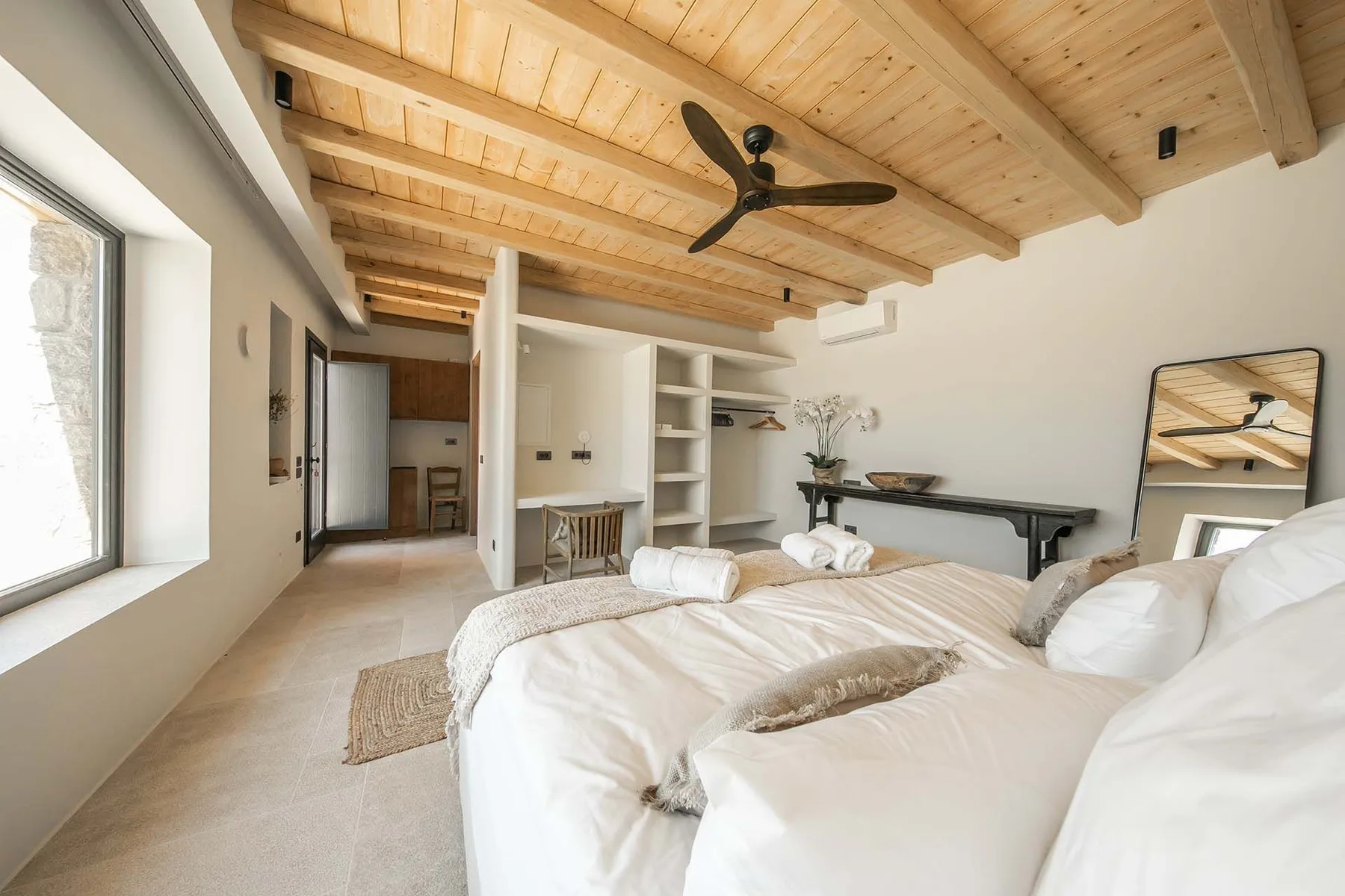 A bright bedroom with white bedding, built-in shelves, and a wood-paneled ceiling.