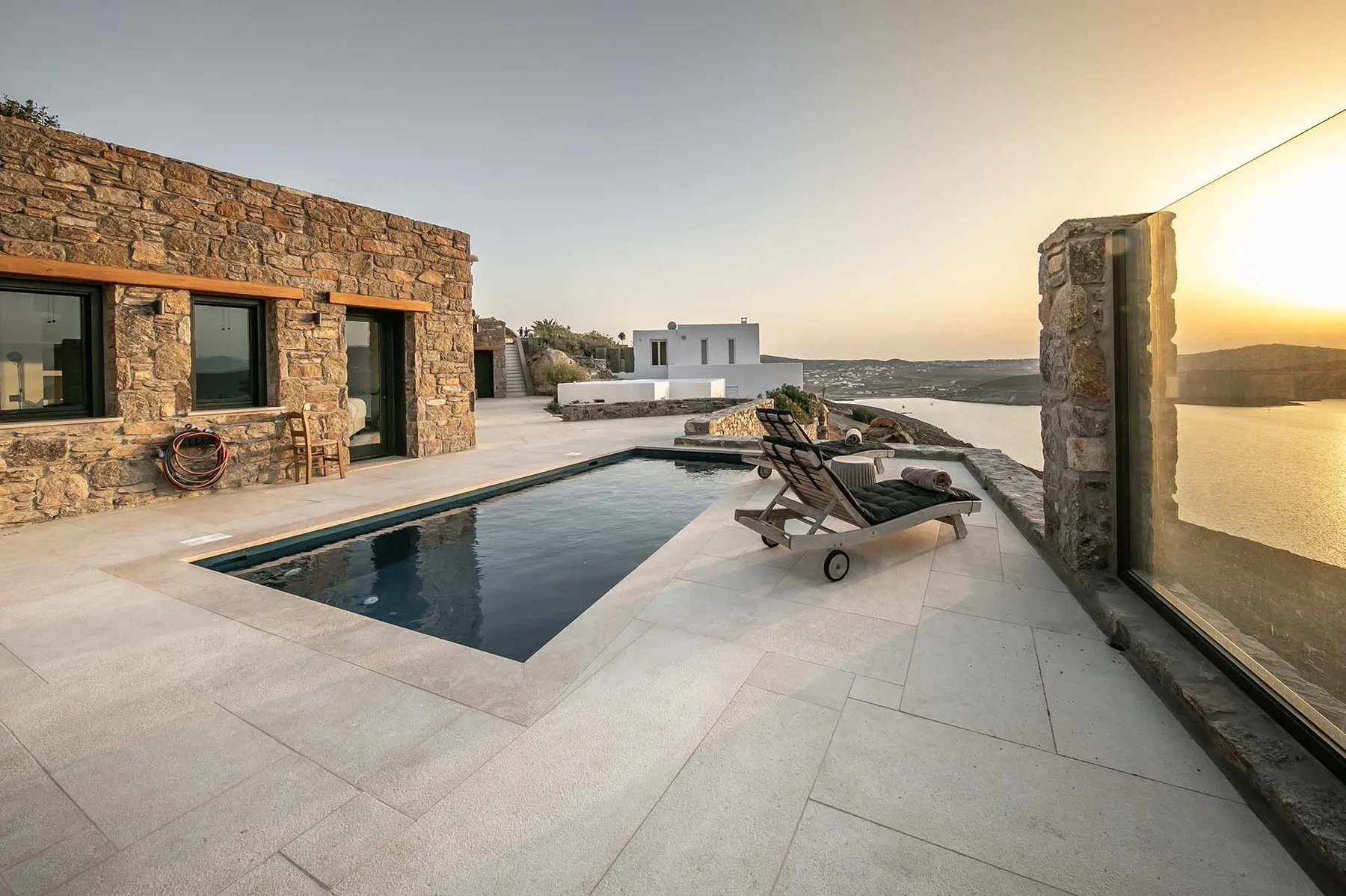 An outdoor pool terrace with lounge chairs beside a stone building at sunset.