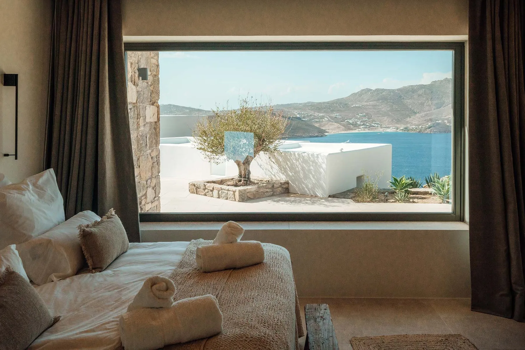 A bedroom with a bed beside a large window overlooking the sea and hills.