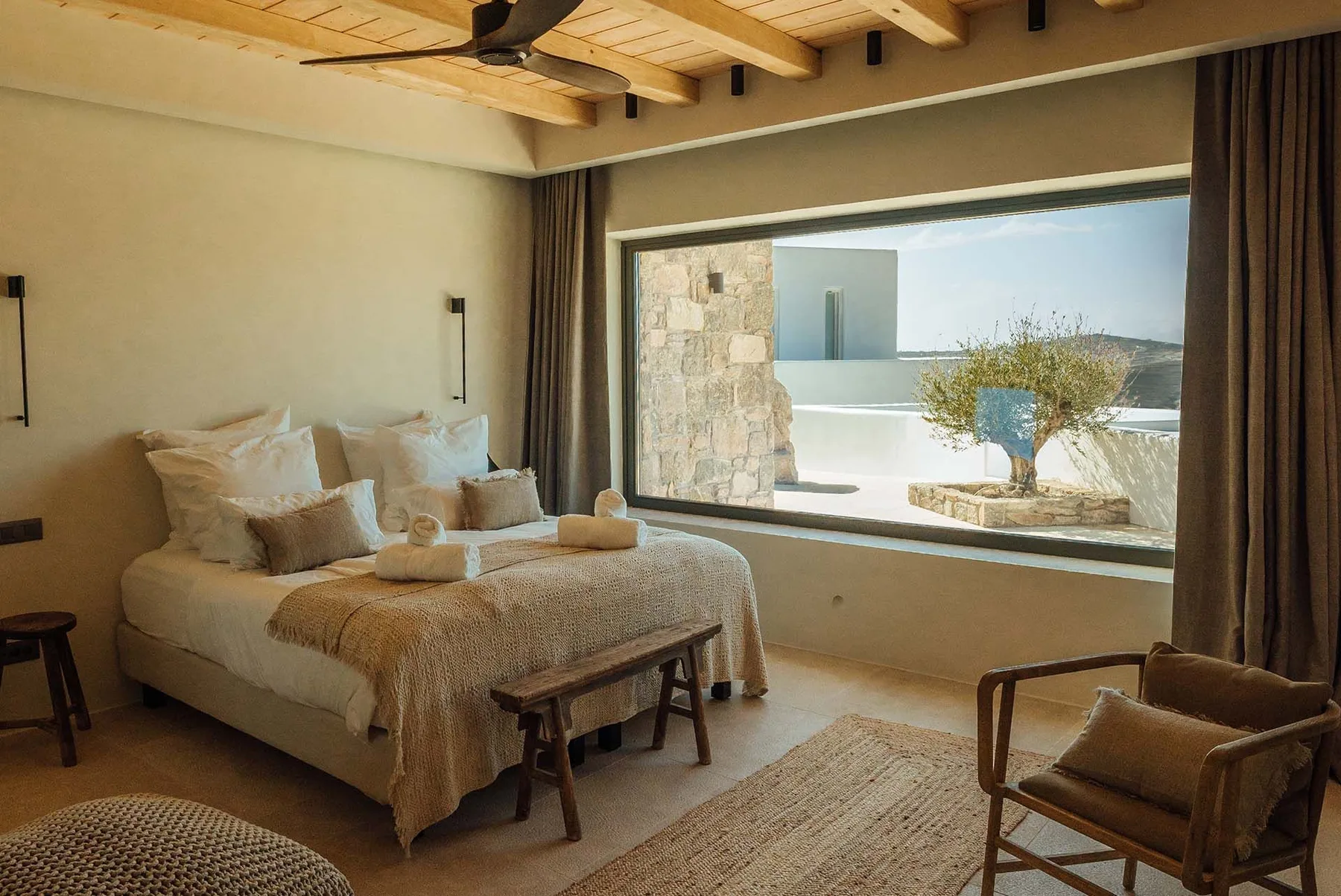 A bedroom with a bed facing a large window overlooking a bright outdoor scene.